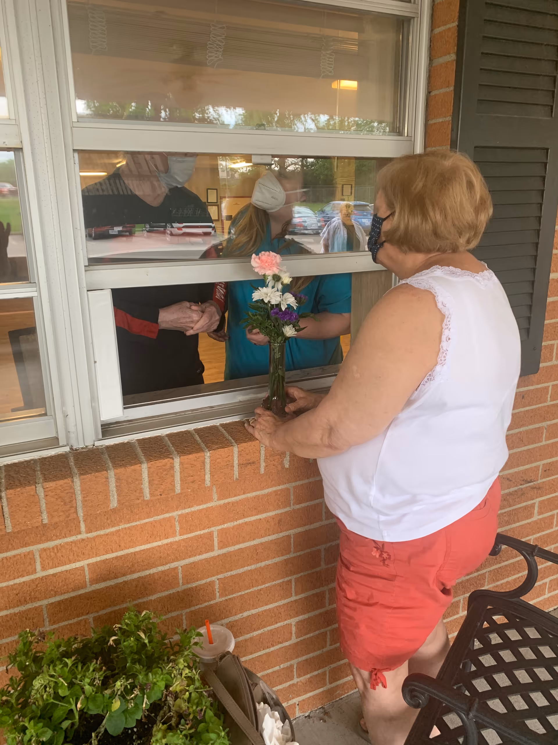 An elderly woman wearing a mask and holding a vase with flowers stands outside a brick building, placing the vase on the windowsill of a partially open window. Inside the building, two masked individuals are visible through the window, one holding the flowers and the other standing nearby. There is a plant and a drink cup on the ground next to the woman.