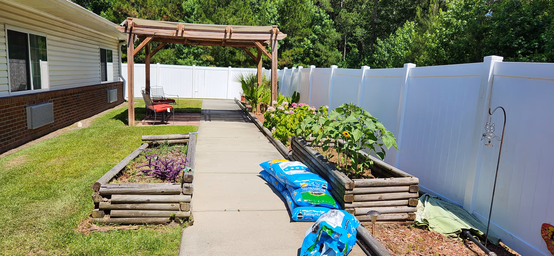 Outdoor garden area at Bolivia Rehabilitation & Healthcare Center featuring a concrete pathway flanked by raised wooden garden beds with various plants and flowers. A wooden pergola with chairs is visible on the left side near the building, and a white privacy fence runs along the right side. Several bags of potting mix are placed on the pathway.
