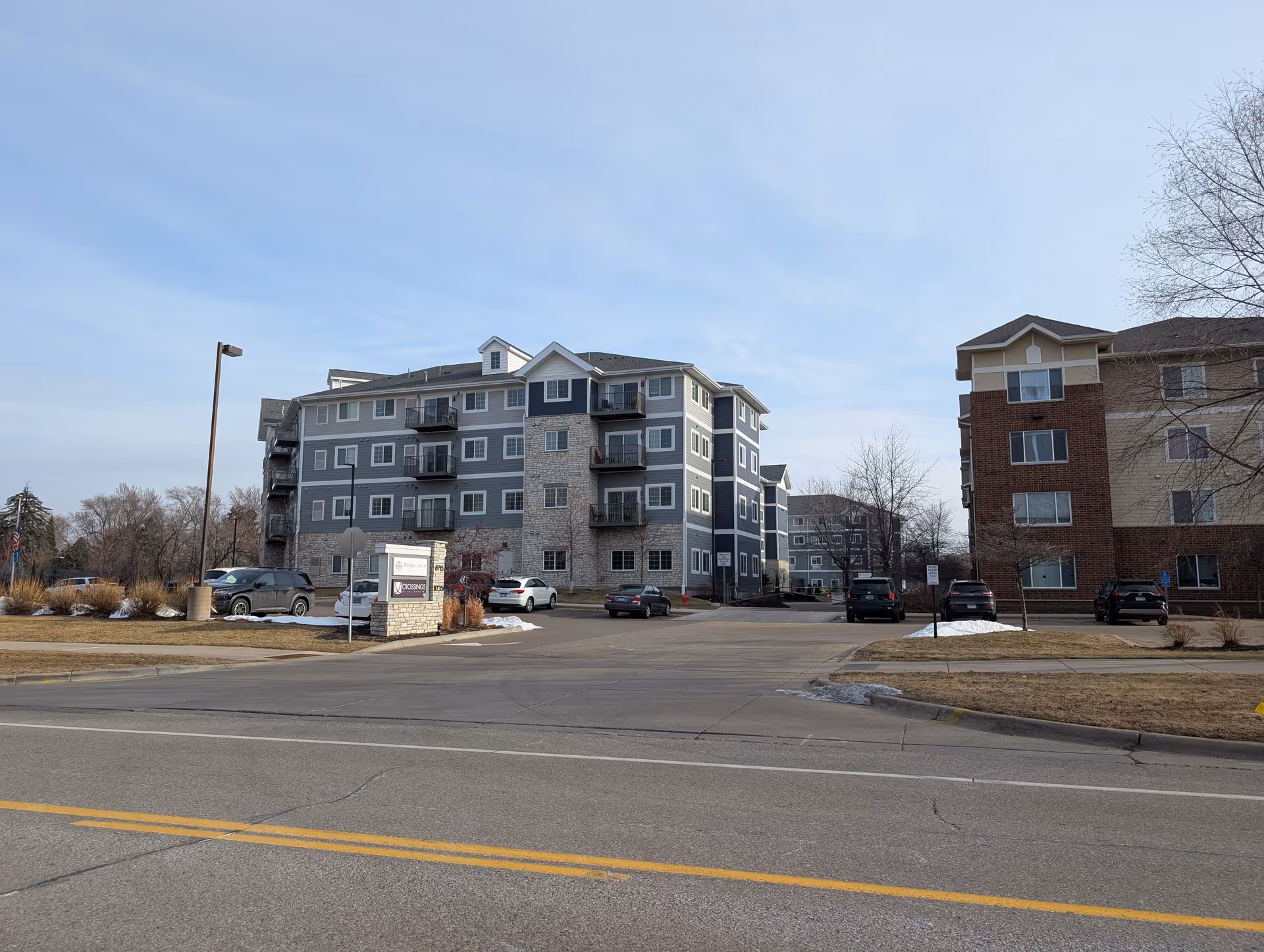 Exterior view of a senior housing facility named Hayden Grove Senior Housing, showing multi-story buildings with balconies, parked cars, and a sign at the entrance. The sky is clear with some light clouds.