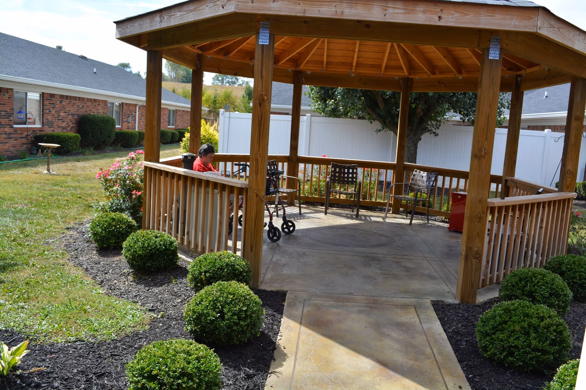 A wooden gazebo in a garden area with neatly trimmed bushes and a concrete pathway leading to it. Inside the gazebo, there are several chairs and a person sitting next to a walker. The background shows a white fence, a tree, and brick buildings.