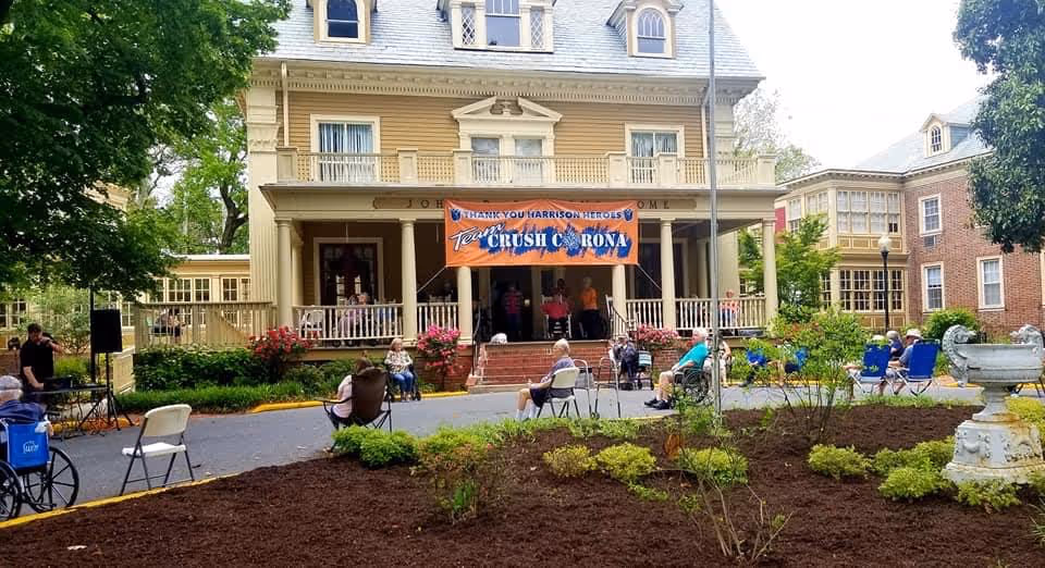 Outdoor gathering at John B. Parsons Assisted Living Community with elderly residents seated in chairs and wheelchairs spaced apart in front of a large, historic-looking building with a porch. A banner on the porch reads 'Thank You Harrison Heroes, Team Crush Corona.' There are plants and trees surrounding the area.