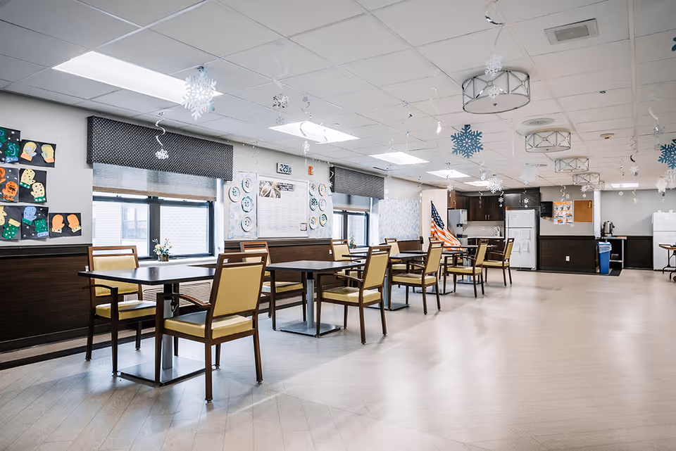 A spacious dining room with several tables and chairs arranged neatly. The room has large windows with black valances, decorated walls with children's artwork and paper plates, and hanging snowflake decorations from the ceiling. An American flag is visible near the back, along with kitchen appliances like refrigerators and a coffee maker.