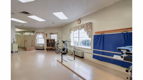 A spacious rehabilitation room with parallel bars for walking exercises, exercise equipment, and large windows with floral curtains allowing natural light. The room has a beige floor and walls, fluorescent ceiling lights, and a blue padded mat on the wall.