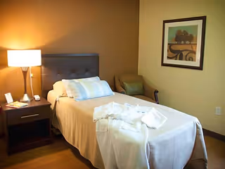 A neatly made single bed with a brown headboard and beige bedding in a cozy room. A bedside table with a lamp, phone, and some items is next to the bed. A comfortable armchair is placed in the corner, and a framed artwork hangs on the wall. The room has warm lighting and a calm atmosphere.