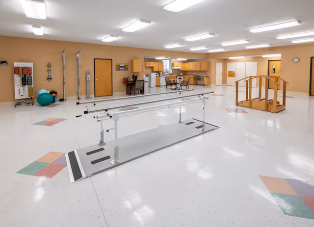 A spacious rehabilitation room with parallel bars for walking exercises in the center, a wooden staircase for physical therapy on the right, and exercise equipment including resistance bands and exercise balls on the left. The room has a light-colored floor with colorful geometric patterns, beige walls, overhead fluorescent lighting, and a kitchenette area with cabinets, a refrigerator, and a table with chairs in the background.