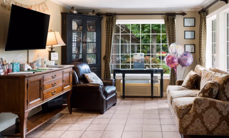 A cozy living room area with a patterned sofa on the right, a dark leather armchair in the center, and a wooden sideboard with a lamp and various items on the left. A flat-screen TV is mounted on the wall above the sideboard. Behind the armchair is a tall black cabinet with glass doors. Large windows with patterned curtains let in natural light and show greenery outside. There are three colorful balloons near the sofa.