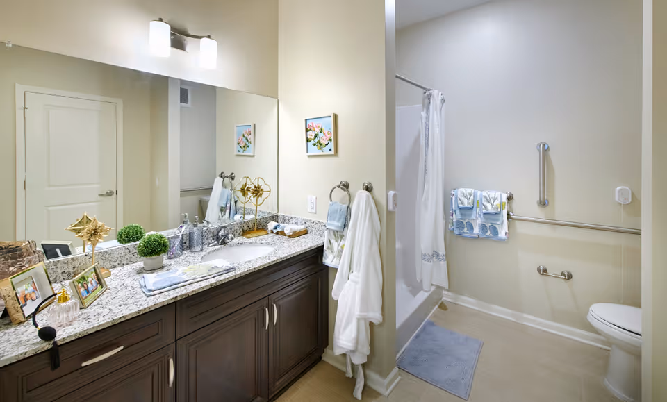 A clean and well-lit bathroom featuring a granite countertop with a sink, decorative items, and framed photos. There is a large mirror above the sink, a white door, a walk-in shower with a white curtain, grab bars on the walls, a towel rack with folded towels, a bathrobe hanging on a hook, and a toilet partially visible on the right side.