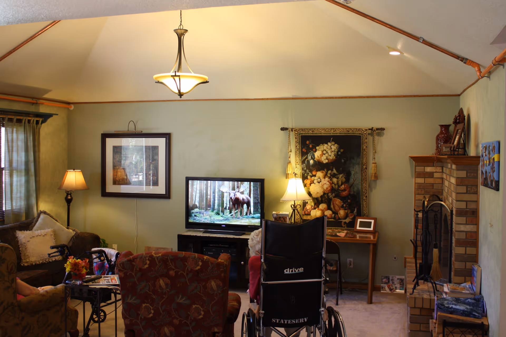A cozy living room with a high vaulted ceiling, featuring a television displaying a moose in a forest, a floral tapestry on the wall, a brick fireplace, and comfortable seating including a wheelchair and armchairs. There are lamps providing warm lighting and various decorative items on tables and shelves.