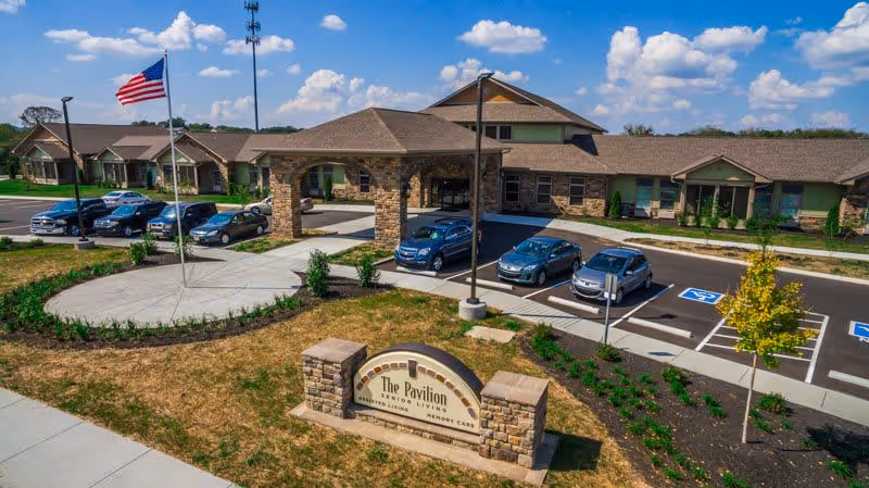 Exterior view of The Pavilion Senior Living at Lebanon facility showing a single-story building with a covered entrance, several parked cars, an American flag on a flagpole, and a landscaped area with a sign displaying the facility's name.
