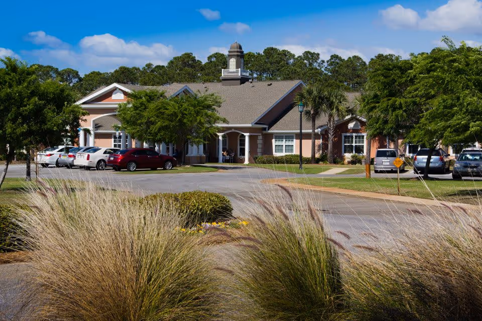 Exterior view of Charter Senior Living of Panama City Beach showing a single-story building with a peaked roof and a small tower on top. The building is surrounded by trees, bushes, and a parking lot with several cars parked. The sky is clear with a few clouds.