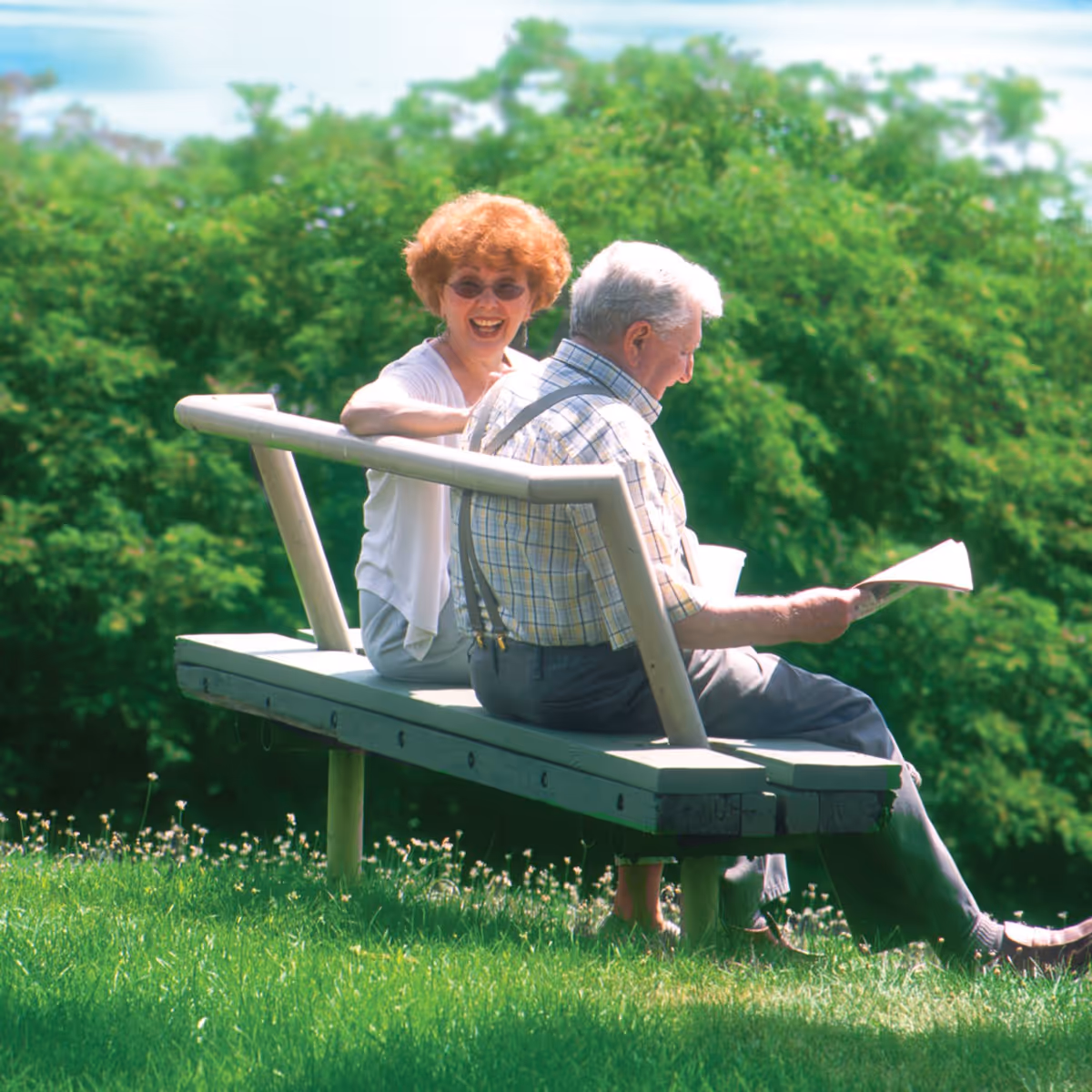 An elderly man and woman sitting on a bench outdoors surrounded by green grass and trees. The man is reading a newspaper while the woman is smiling and looking towards the camera.