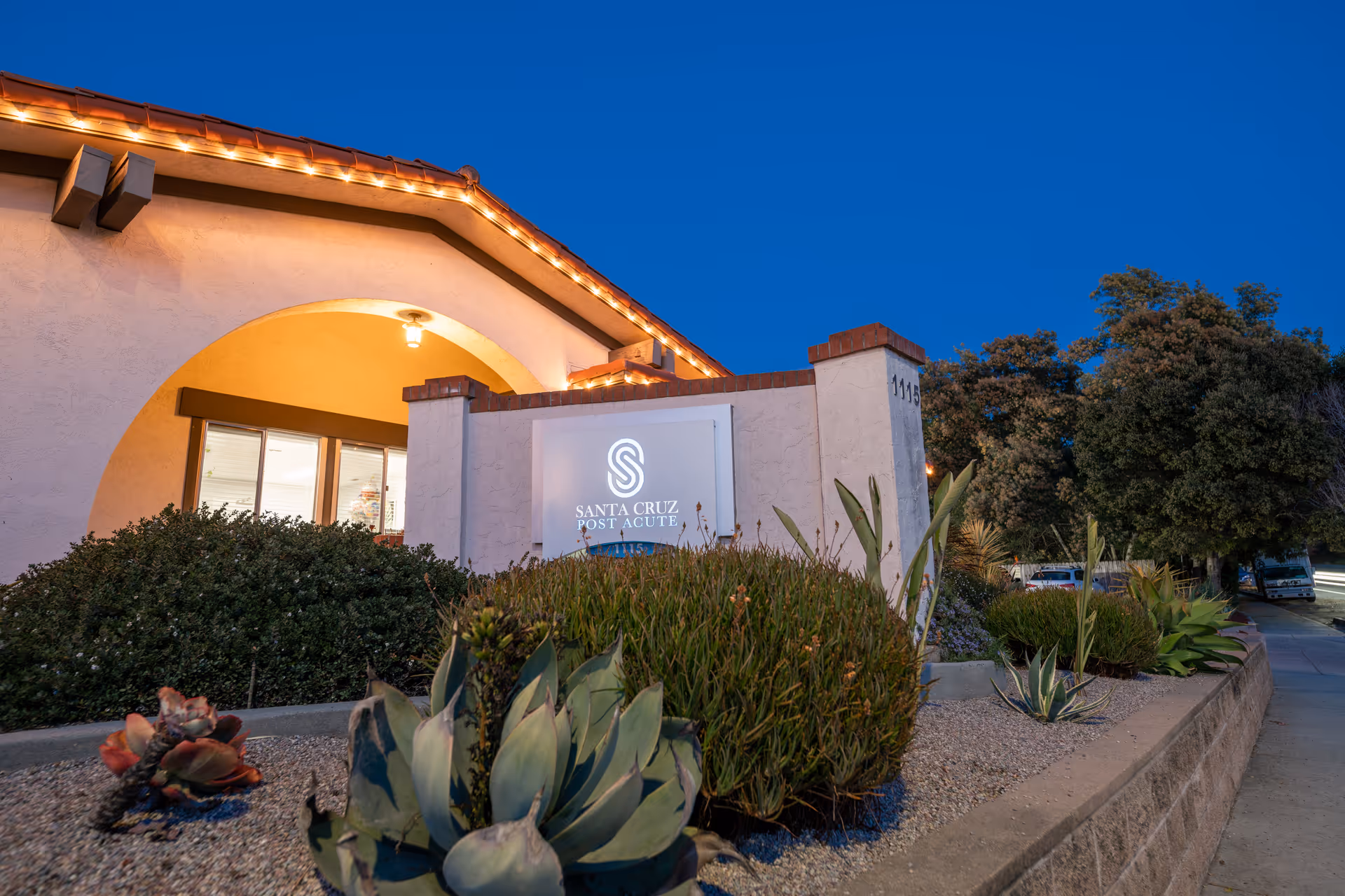 Exterior front entrance of Santa Cruz Post Acute building with illuminated sign and landscaped succulents at dusk.