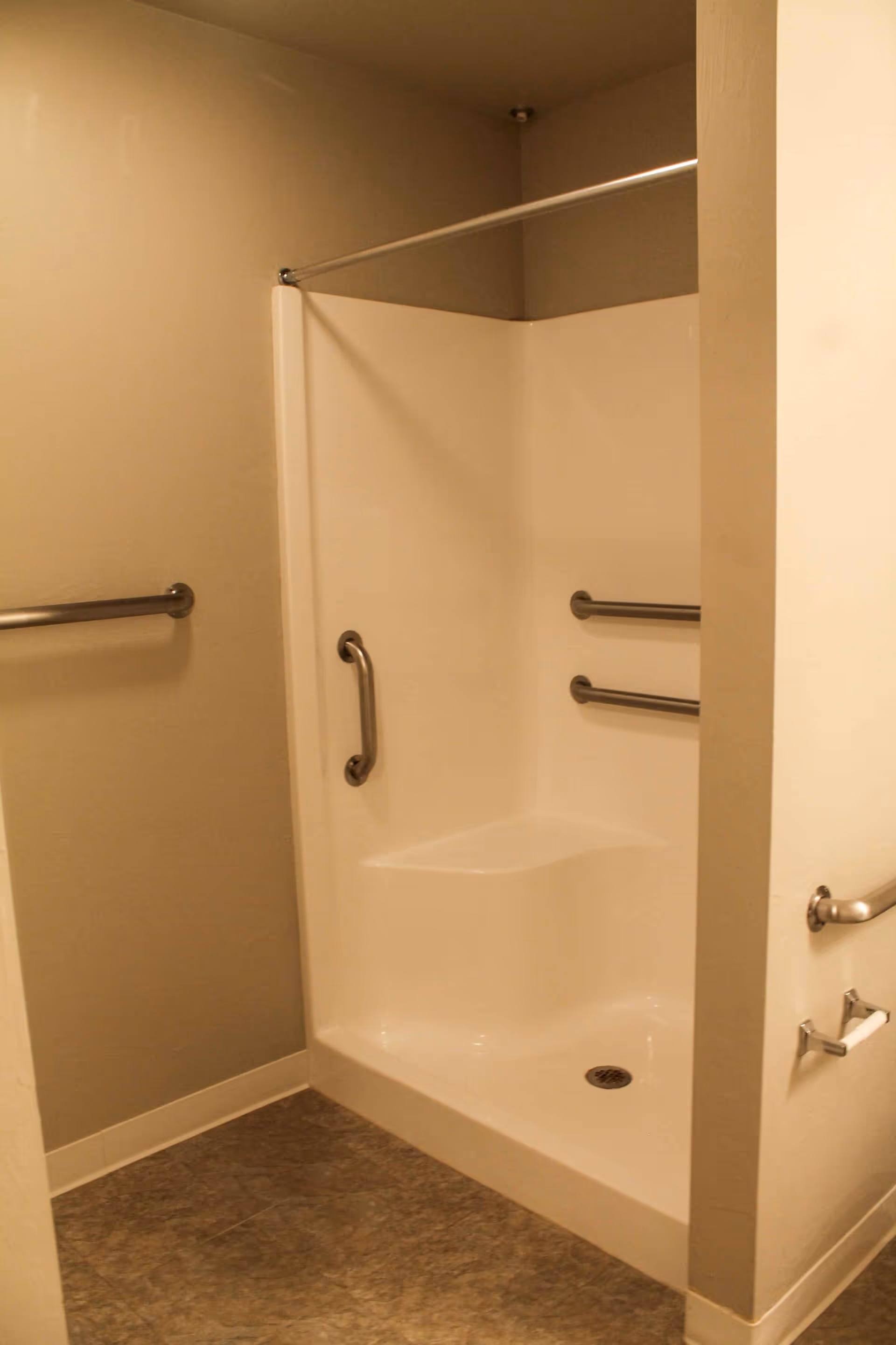 A beige bathroom with a walk-in shower featuring built-in seating and multiple stainless steel grab bars for support. The floor has a textured pattern, and there is a towel bar and a toilet paper holder mounted on the walls.