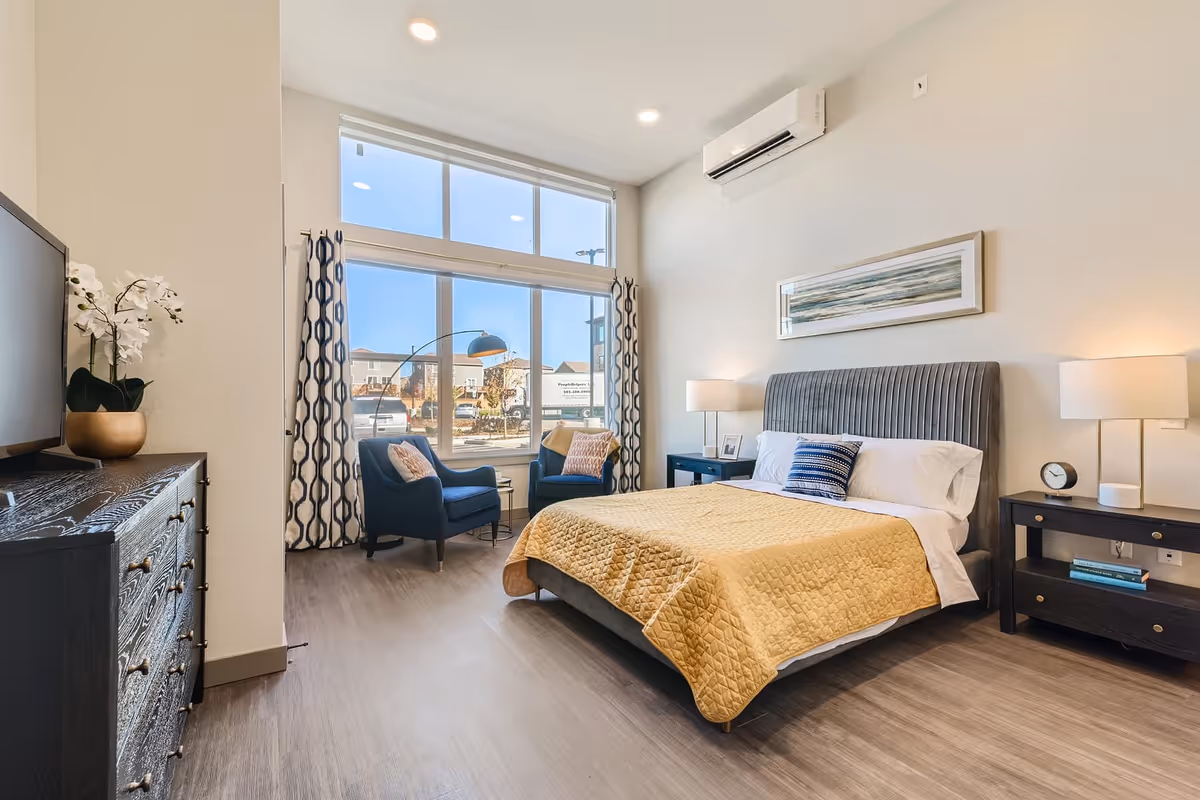 A bright and modern bedroom with a large window letting in natural light. The room features a bed with a yellow quilt and blue and white pillows, two dark wood nightstands with lamps, a dark wood dresser with a TV on top, and two blue armchairs near the window with patterned curtains. The floor is light wood, and there is a wall-mounted air conditioning unit above the bed.