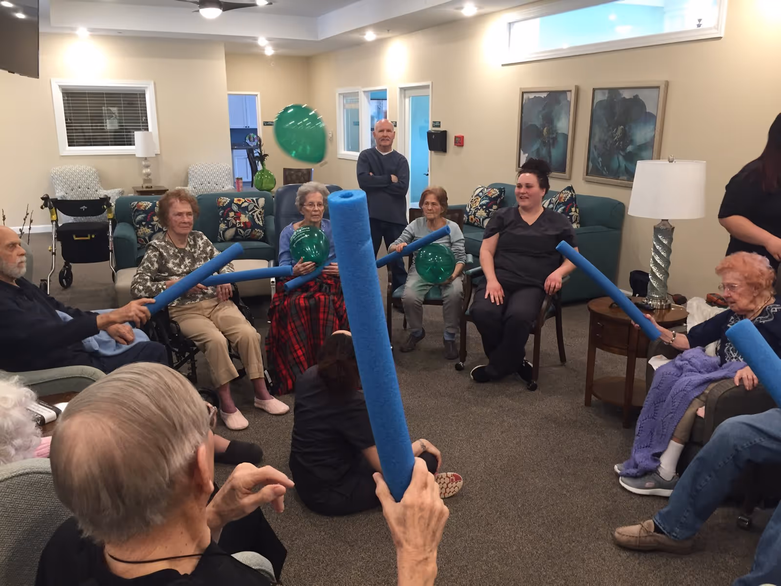 A group of elderly people and caregivers sitting in a circle in a living room, engaging in a balloon exercise activity using blue foam noodles. The room is furnished with chairs, couches, and decorated with floral paintings and lamps.