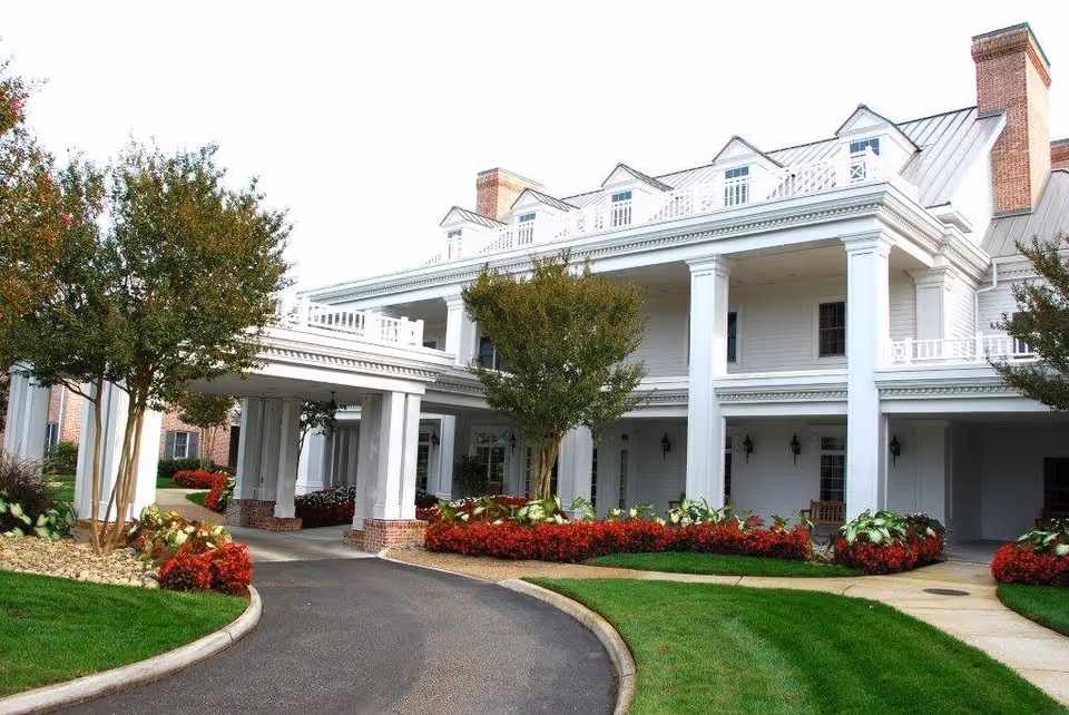 Front exterior view of a large white retirement and assisted living community building with tall columns, manicured green lawns, red flower beds, and trees along a curved driveway.
