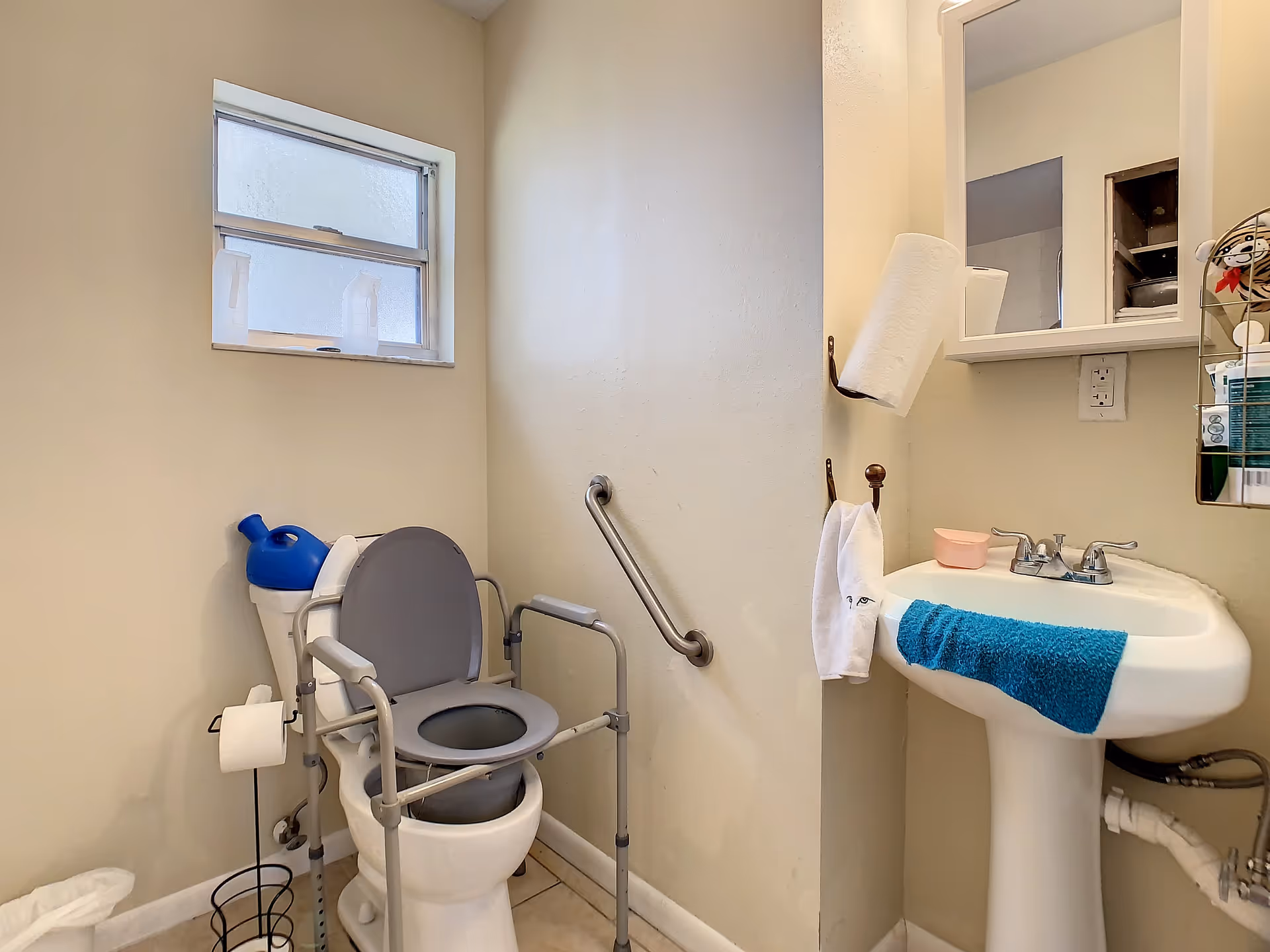 Small bathroom with a toilet equipped with a gray raised seat and support frame, a grab bar on the wall, a pedestal sink with a blue towel draped over the front, a paper towel roll holder mounted on the wall, and a small frosted window above the toilet.
