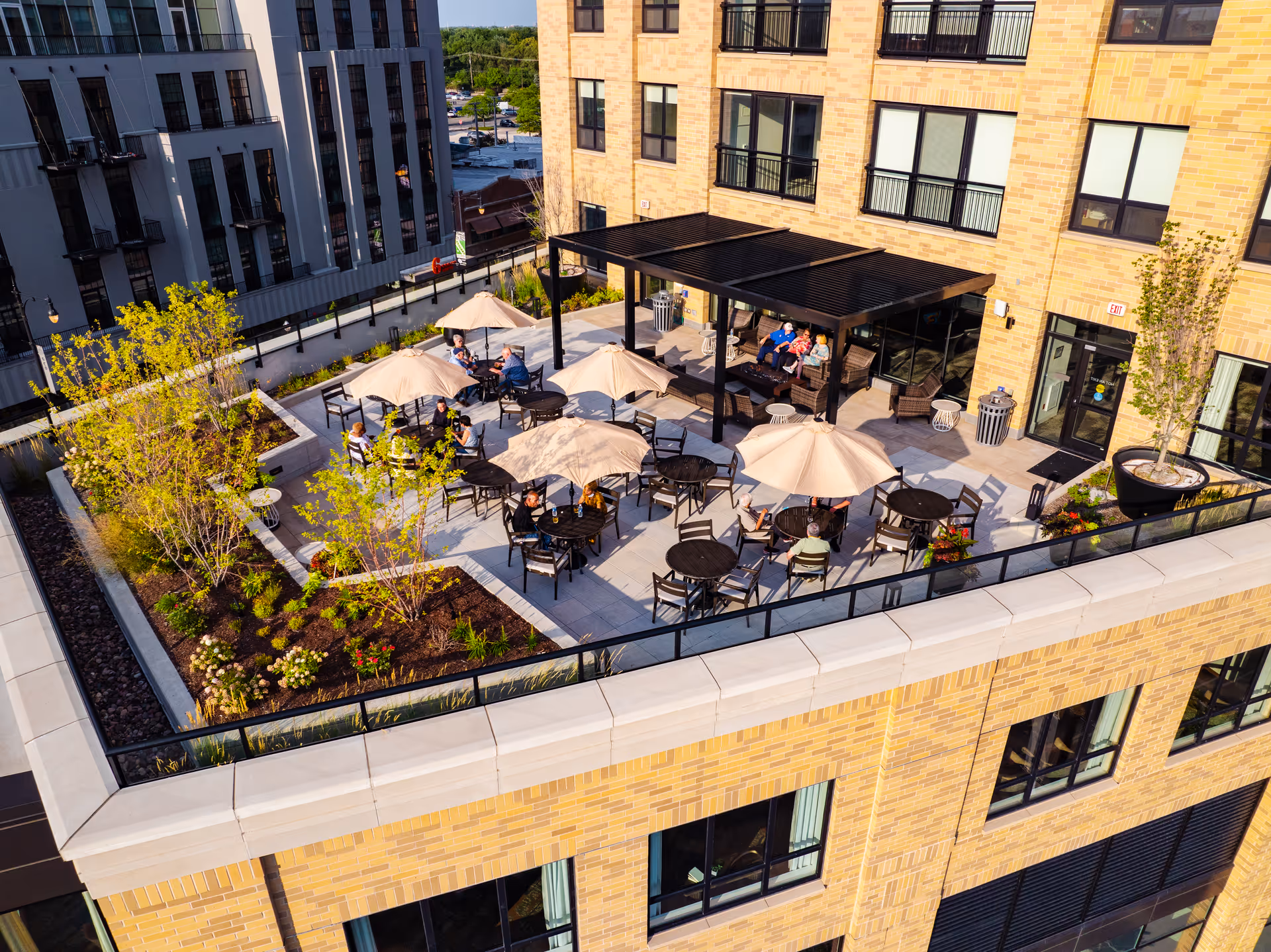 Rooftop outdoor patio with tables, umbrellas, planters and people seated outside a multi-story brick senior living building.
