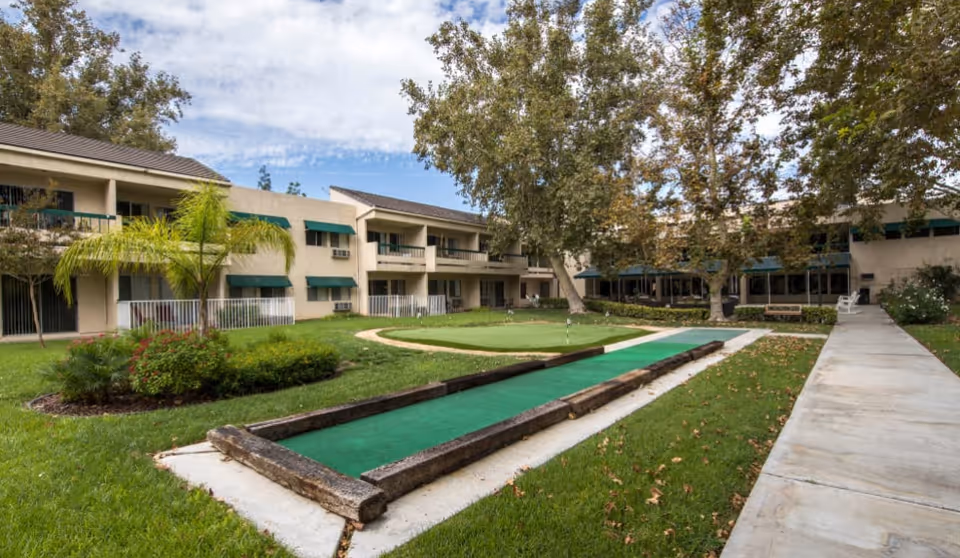 Outdoor courtyard of a two-story senior living building featuring a small putting green, walkways, and landscaped trees.