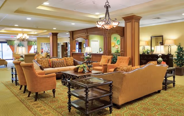A spacious senior living facility common area with comfortable upholstered sofas and armchairs arranged around a wooden coffee table. The room features warm lighting from table lamps and a chandelier, decorative plants, and a reception desk in the background with wood paneling and colorful wall decor.