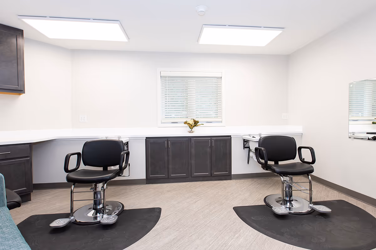 A bright, clean salon room with two black salon chairs on black mats facing a wall with a window and dark cabinetry underneath. The room has light-colored walls and flooring, with two large ceiling lights providing illumination.