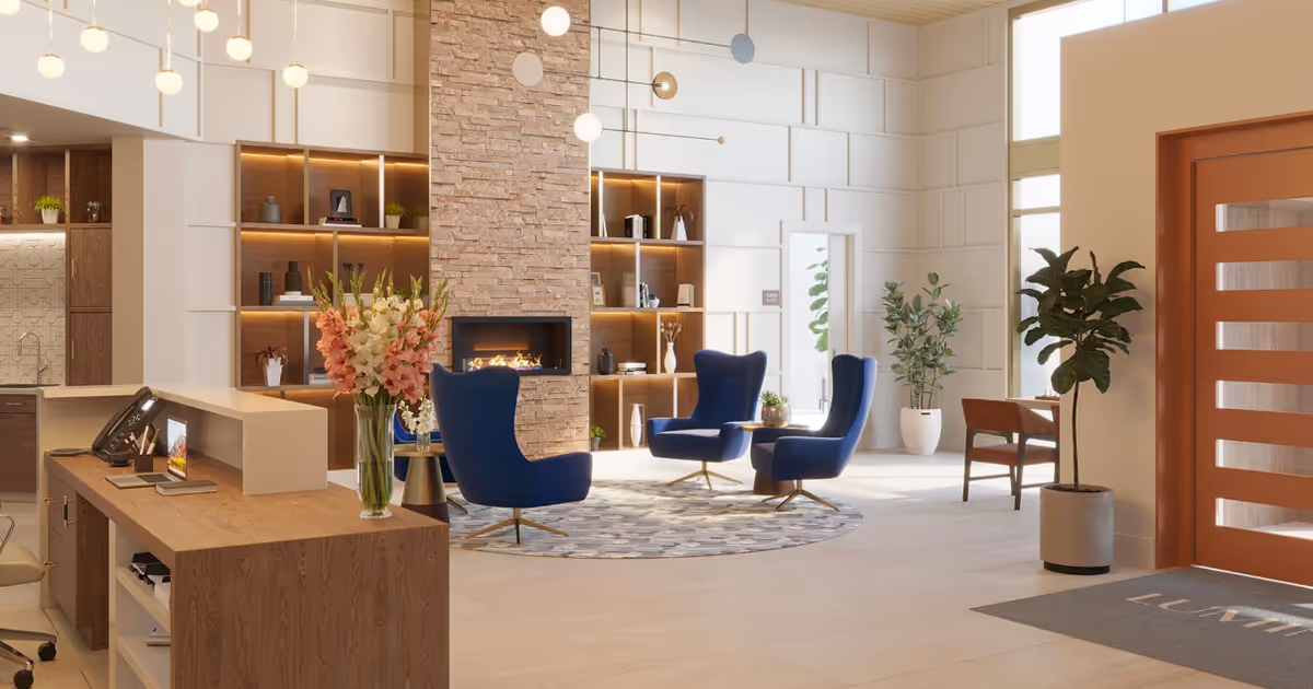 A modern and bright senior living facility lobby with a wooden reception desk on the left, a vase of flowers on the desk, and a seating area featuring four blue armchairs arranged around a circular rug in front of a stone fireplace. The walls have built-in shelves with decorative items and plants. Large windows allow natural light to fill the space, and there are additional plants and a wooden door on the right with a mat that reads 'LUMINA'.