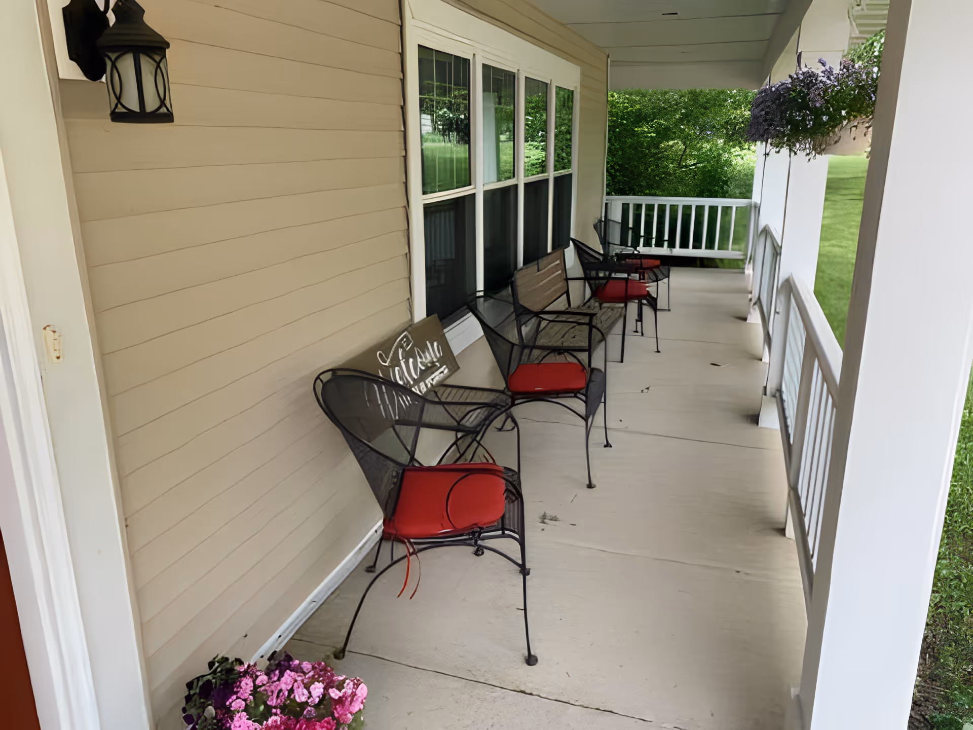 Covered porch area with beige siding, four black metal chairs with red cushions, a bench with a decorative sign, hanging flower baskets, and a view of green grass and trees.