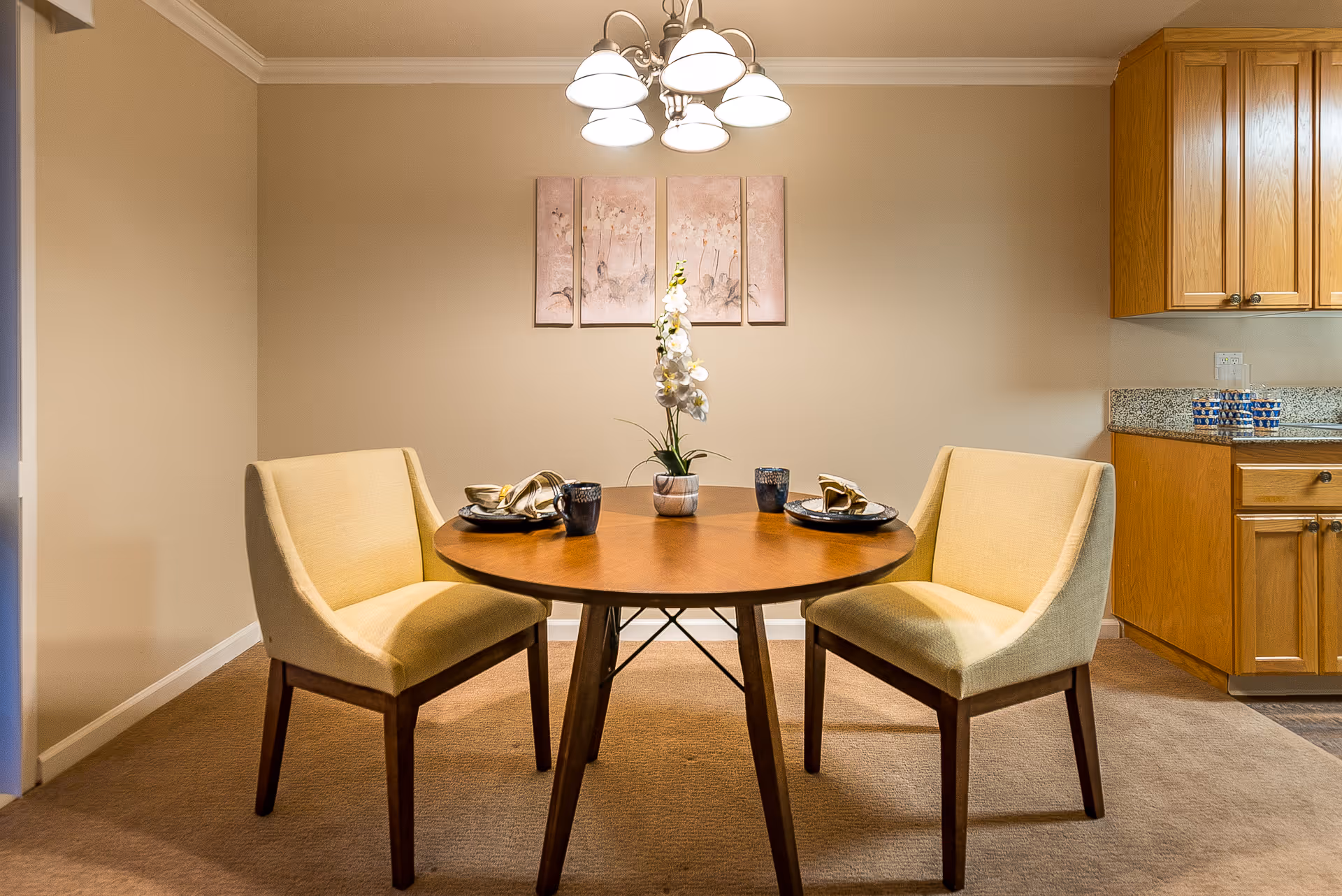 A small dining area with a round wooden table set for two with plates, napkins, and mugs. Two beige upholstered chairs are positioned on either side of the table. A white orchid centerpiece is on the table. Above the table hangs a chandelier with five lights. On the wall behind the table are four vertical art panels with floral designs. To the right, part of a kitchen with wooden cabinets and granite countertops is visible.