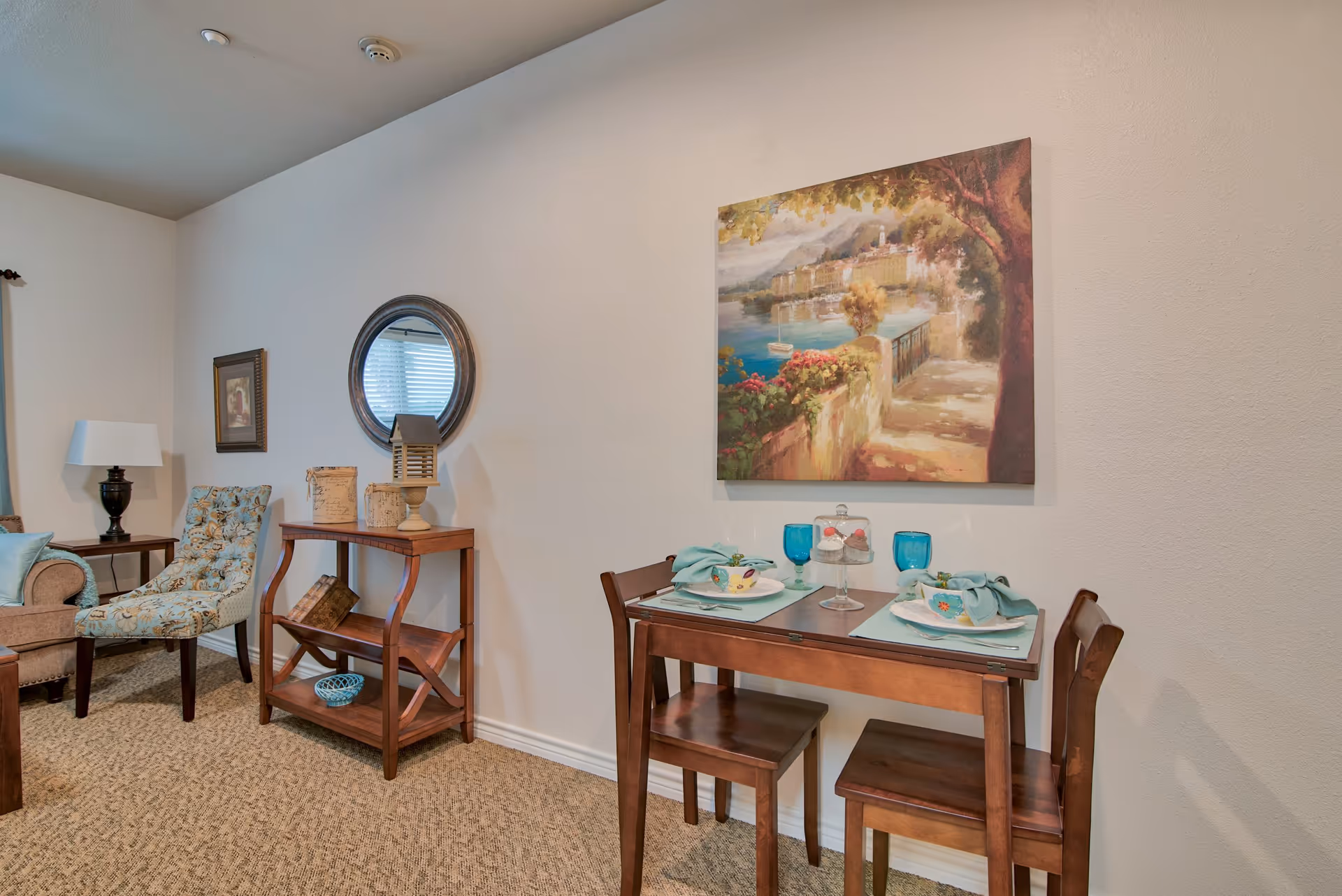 A cozy interior corner of a senior living facility featuring a small wooden dining table set for two with blue glasses and napkins, a decorative painting of a scenic waterfront path on the wall above, a wooden console table with decorative items and books, a round mirror, a floral upholstered chair, and a beige armchair with a lamp on a side table.
