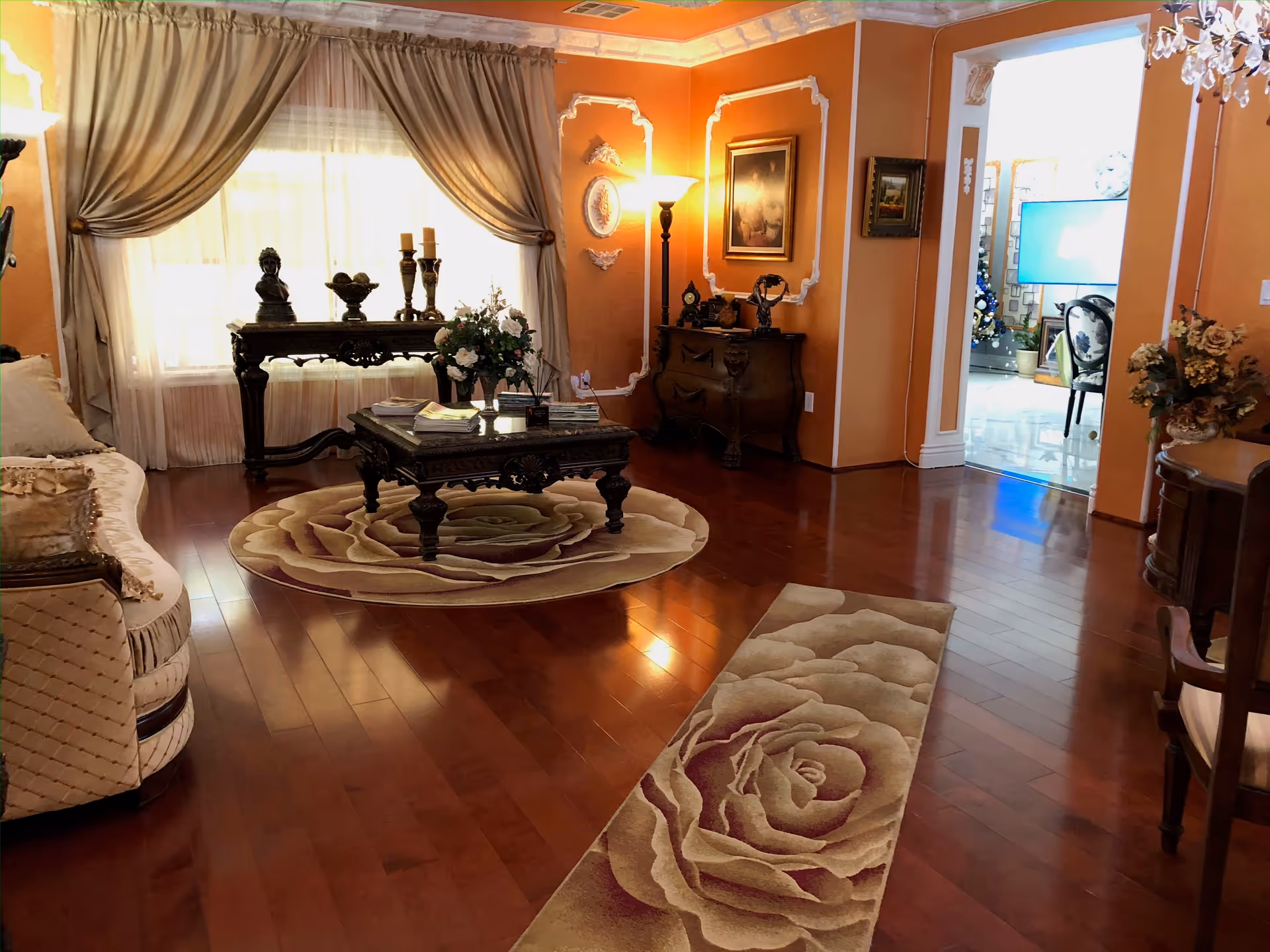 A warmly lit living room with polished wooden floors, featuring a beige sofa with decorative pillows on the left, two floral-patterned rugs on the floor, a dark wooden coffee table with books and a flower arrangement, and a matching console table with decorative items near a window with beige curtains. The walls are painted orange with white decorative moldings, and there are framed paintings and a floor lamp. An adjoining room with a TV and chairs is visible through an open doorway.