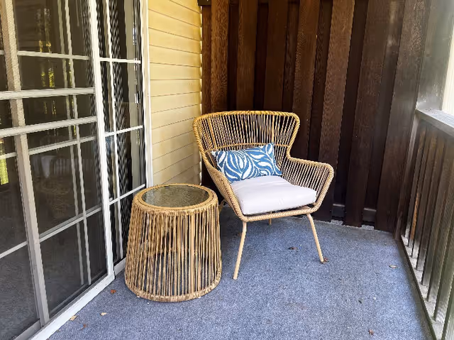 A small outdoor balcony area with a wicker chair featuring a white cushion and a blue patterned pillow, next to a matching wicker side table with a glass top. The balcony has a gray carpeted floor, wooden railing, and sliding glass doors leading inside.
