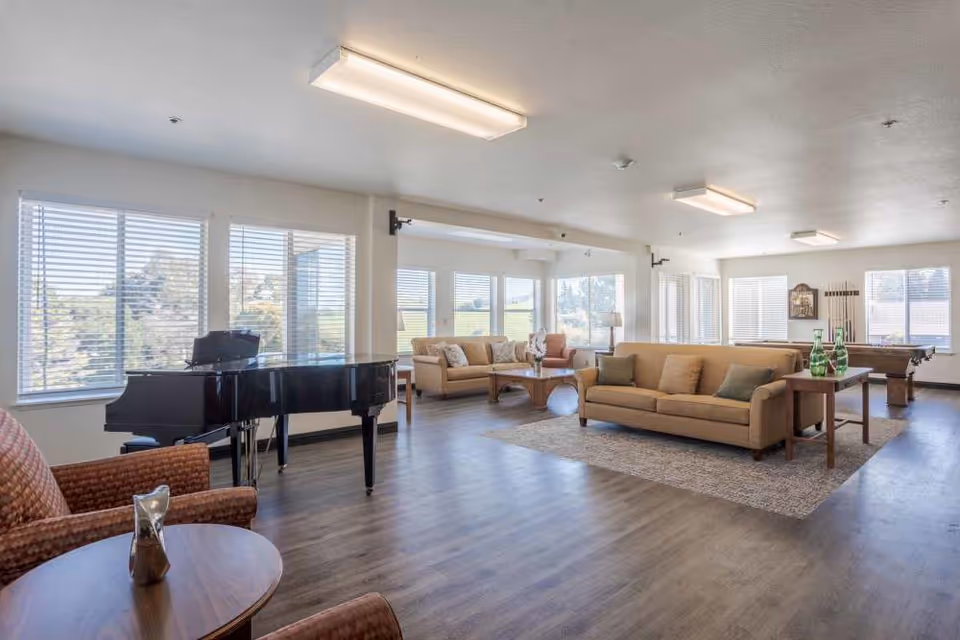 A spacious and well-lit living room area with large windows allowing natural light to fill the space. The room features a black grand piano near the windows, two beige sofas with cushions, a wooden coffee table, and a side table with decorative green glass bottles. There is also a pool table in the background and a patterned rug on the floor. The flooring is wood-style, and the ceiling has rectangular fluorescent light fixtures.