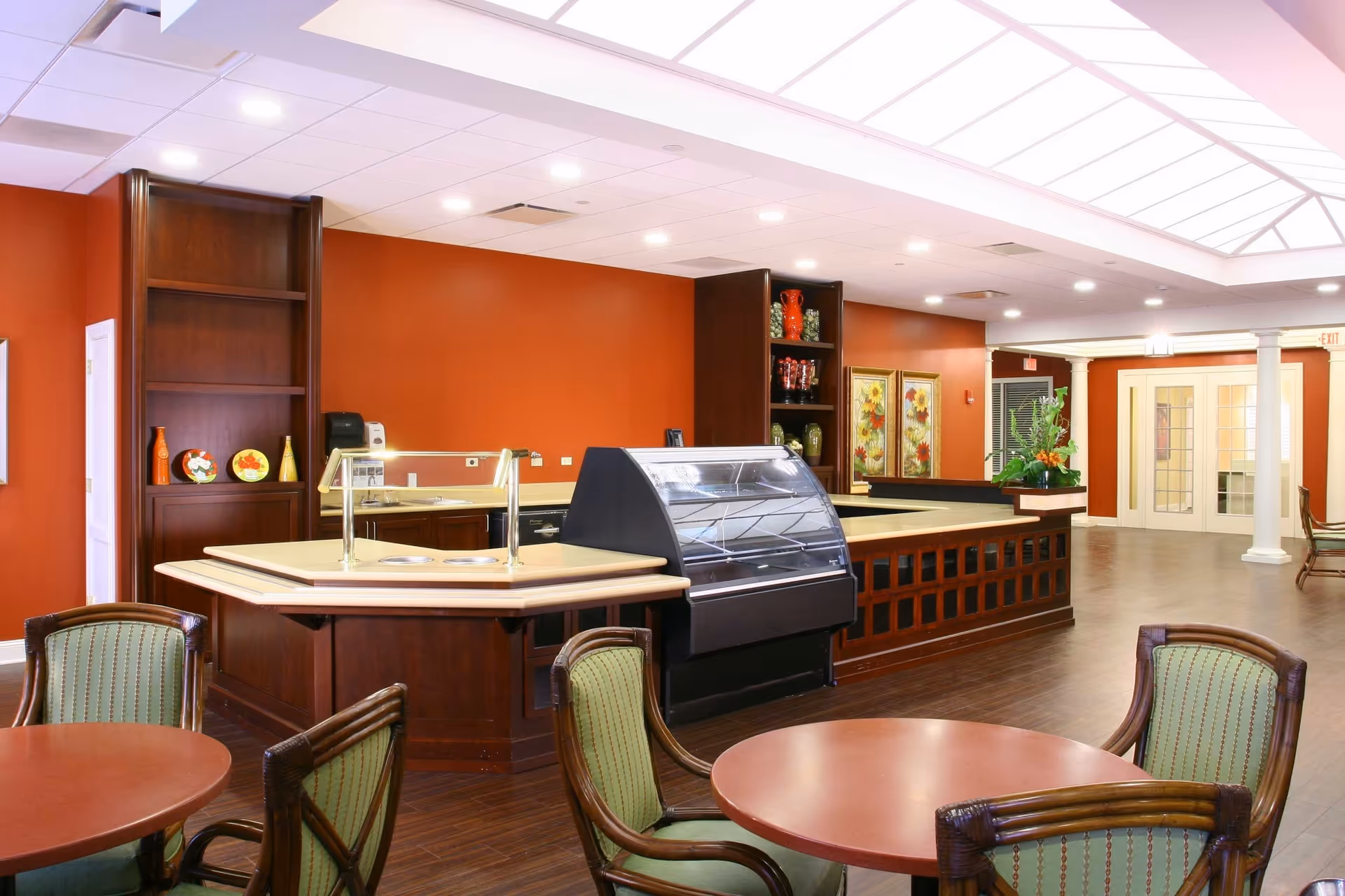 Interior view of a senior living facility dining area with round tables and green cushioned chairs. There is a serving counter with a glass display case and wooden cabinetry against a warm orange wall. The ceiling has recessed lighting and a large skylight panel.