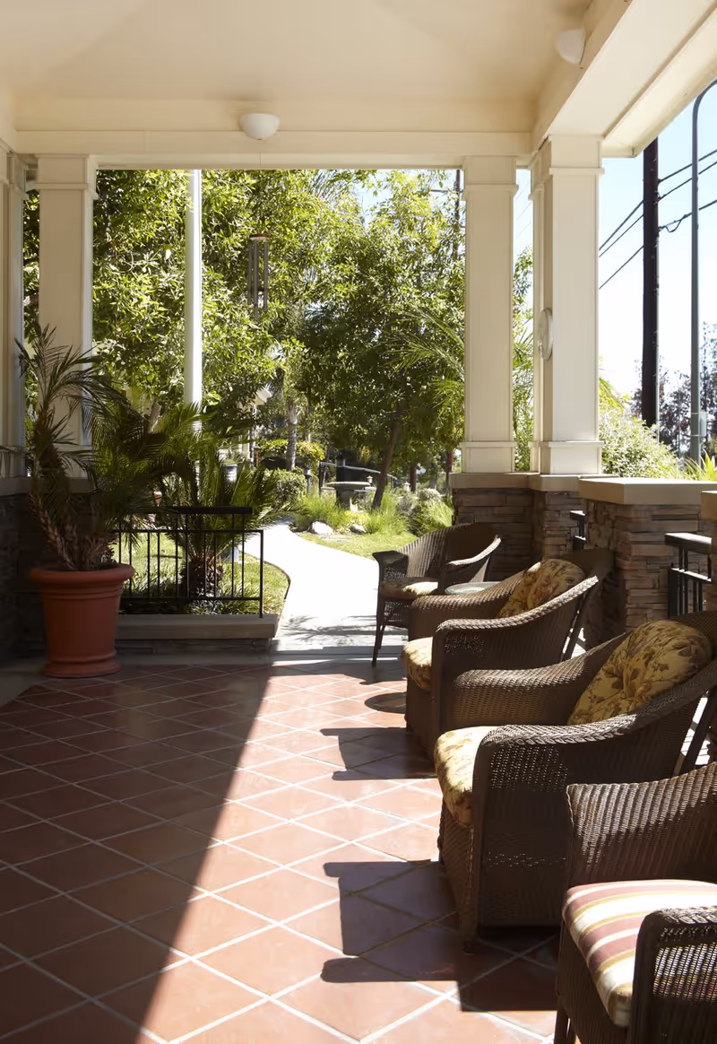 A covered outdoor patio area with wicker chairs featuring floral and striped cushions. The patio has terracotta tile flooring and is surrounded by white pillars and stone accents. Green trees and plants are visible beyond the patio, along a paved walkway.
