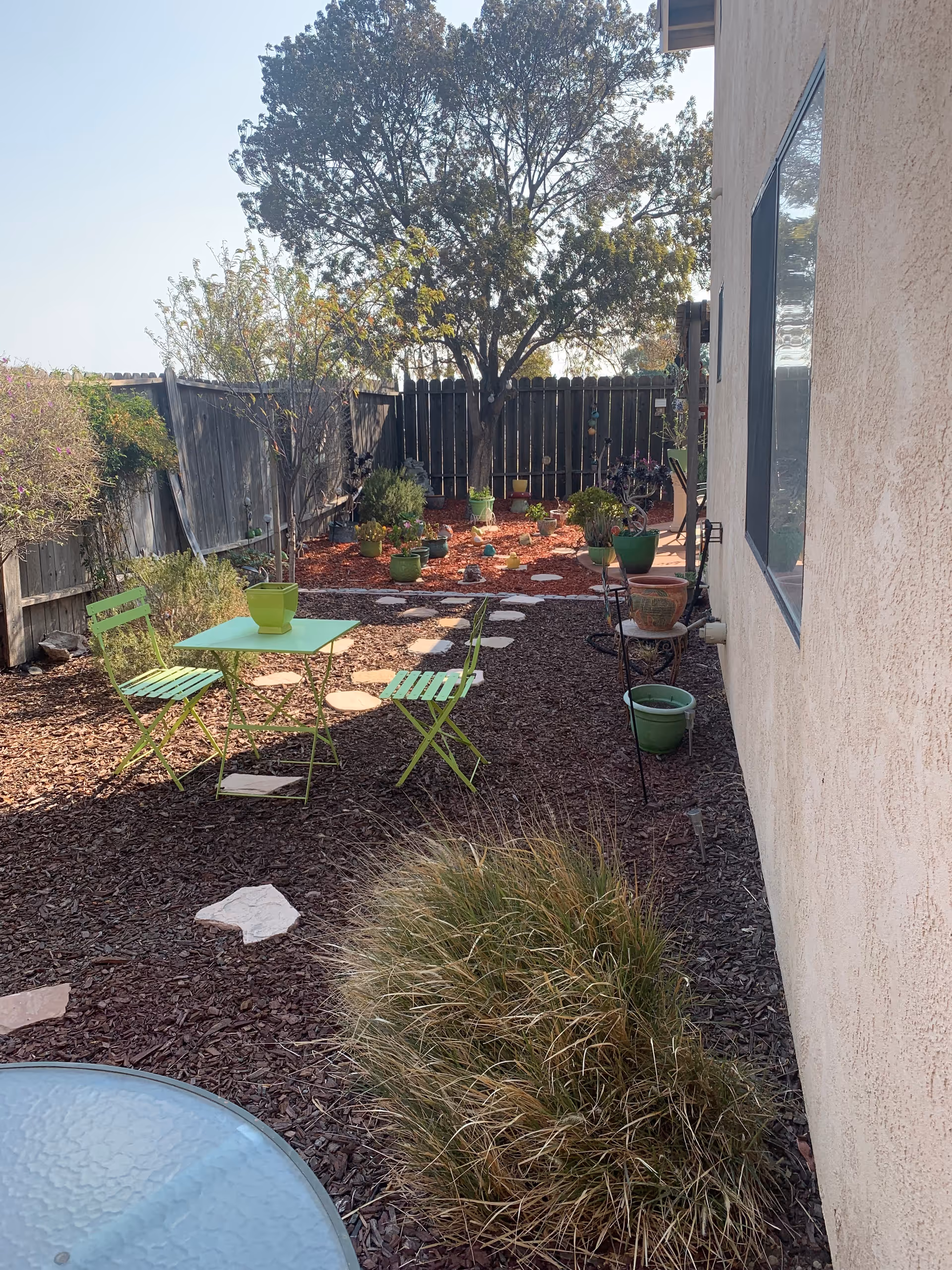Outdoor garden area with a small green metal table and two matching chairs on a mulch-covered ground. There are various potted plants and a tree in the background, with a wooden fence enclosing the space. A beige building wall with a window is visible on the right side.
