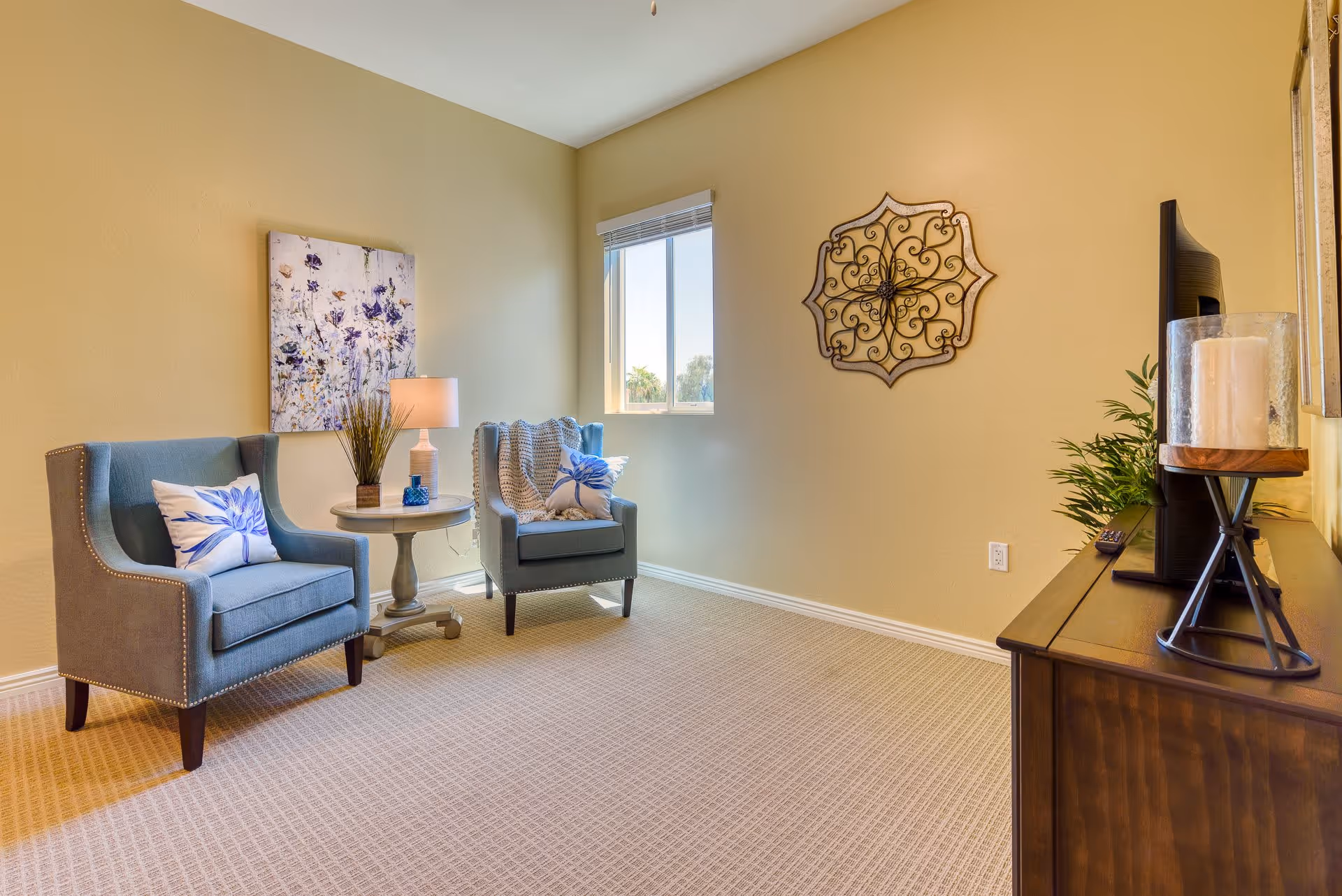 A cozy living room area with two blue armchairs featuring floral pillows, a small round table with a lamp and decorative items between them, a window letting in natural light, a decorative wall piece, and a wooden cabinet with a TV and candle on top.