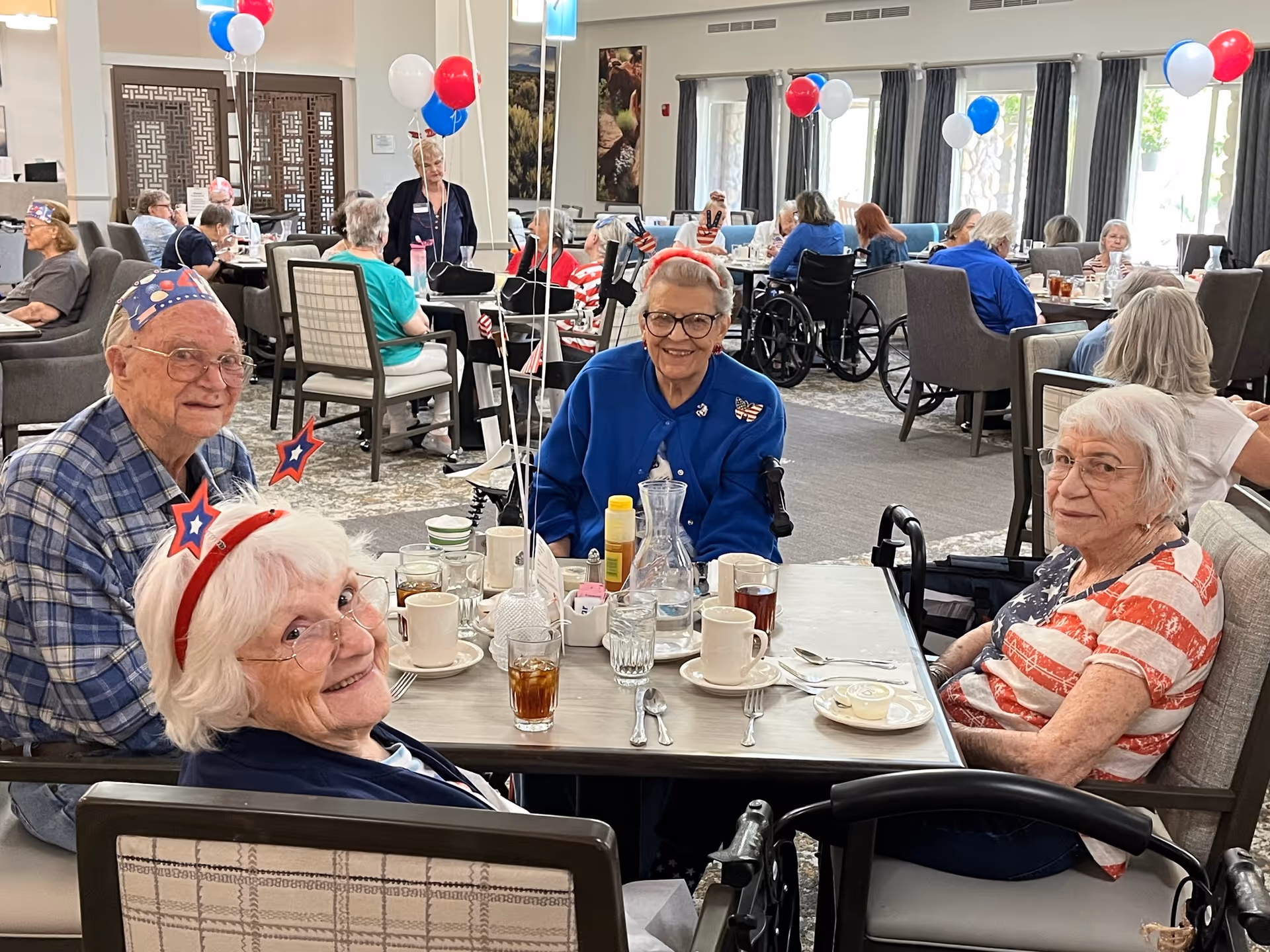 A group of elderly people sitting around a dining table in a decorated dining room, wearing patriotic accessories like star headbands and hats. The room is filled with other seniors seated at tables, some in wheelchairs, with red, white, and blue balloons tied to chairs and festive decorations visible.