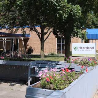 Outdoor view of Parkview Care Center of Heartland Senior Living showing a brick building with a blue roof, a tree, flower beds with pink and red flowers, and a sign with the Heartland Senior Living logo and the facility name.