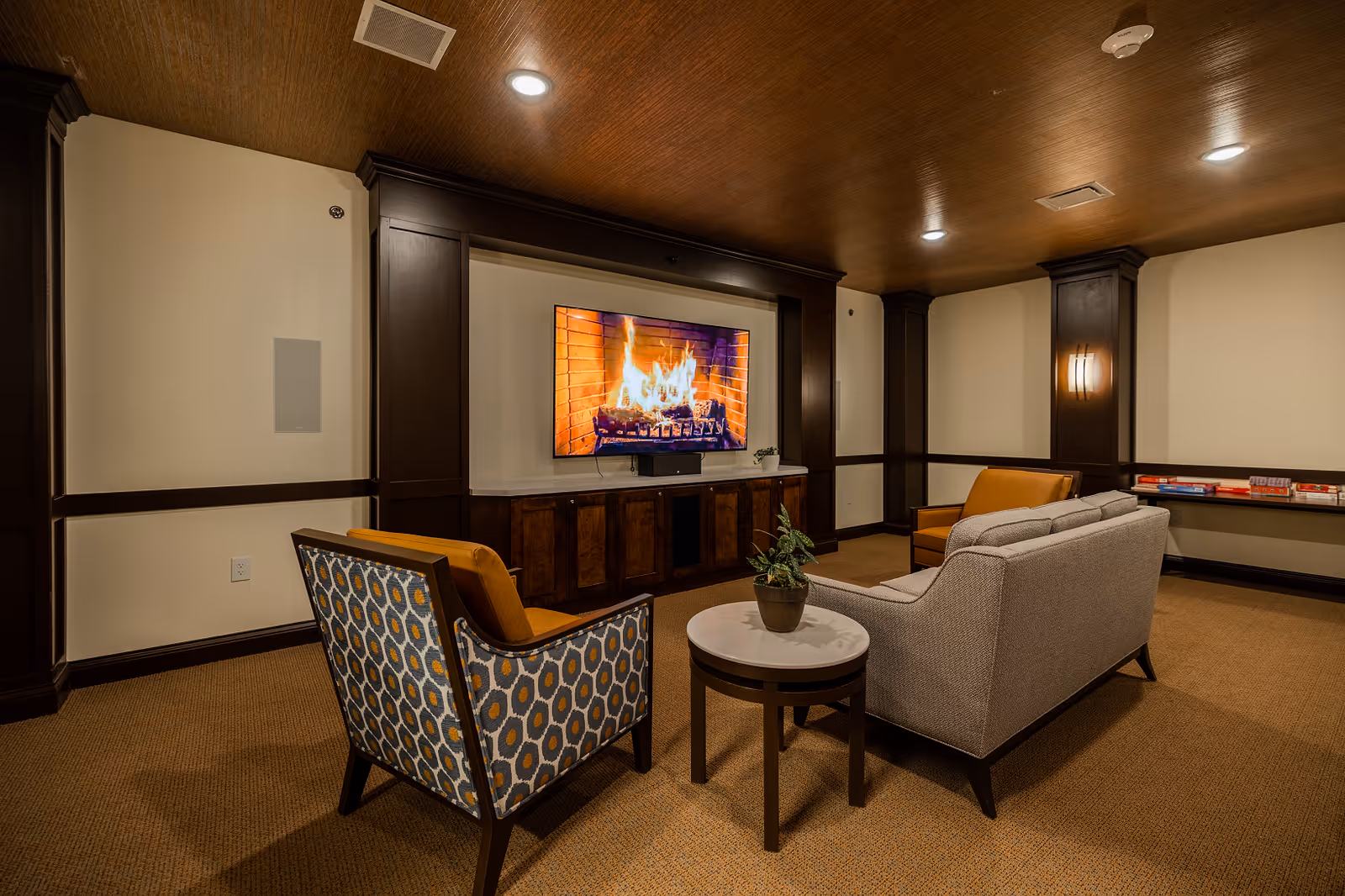 A cozy lounge area with a patterned armchair, a gray sofa, and a mustard yellow armchair arranged around a small round table with a potted plant. A large flat-screen TV mounted on the wall displays a burning fireplace. The room has warm lighting, wood-paneled ceiling, and beige walls with dark wood trim.