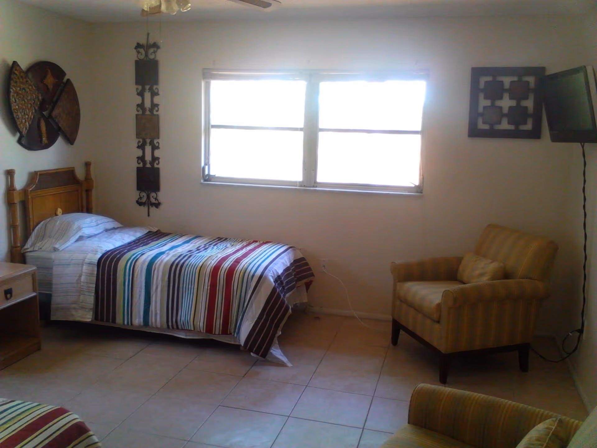 A simple bedroom with a single bed covered in a colorful striped blanket, a wooden headboard, a nightstand, a window letting in bright light, a wall-mounted TV, and two striped armchairs.