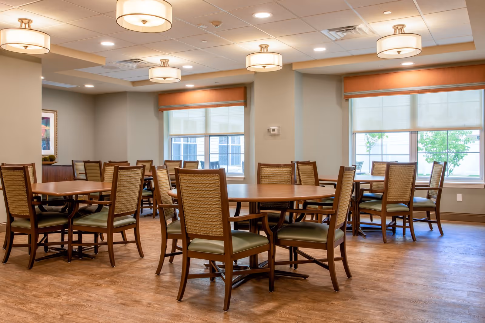 Bright communal dining room with round wooden tables and chairs beneath ceiling light fixtures.