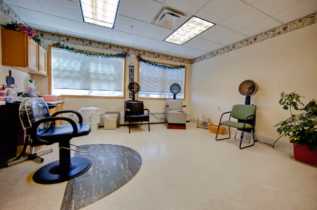 A bright room with large windows covered by blinds, featuring salon chairs and hair drying stations. The room has light-colored walls with floral border wallpaper near the ceiling, a few chairs arranged along the walls, and some plants in the corner.