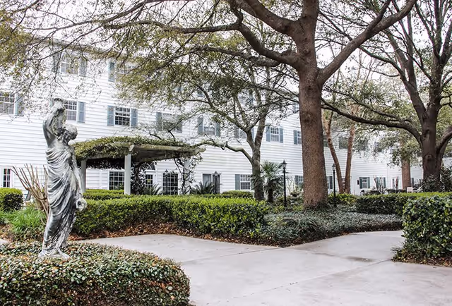 Outdoor view of The Palms of Mt. Pleasant facility showing a white multi-story building with multiple windows, surrounded by large trees, trimmed hedges, and a concrete pathway. A classical statue is visible on the left side near the pathway.