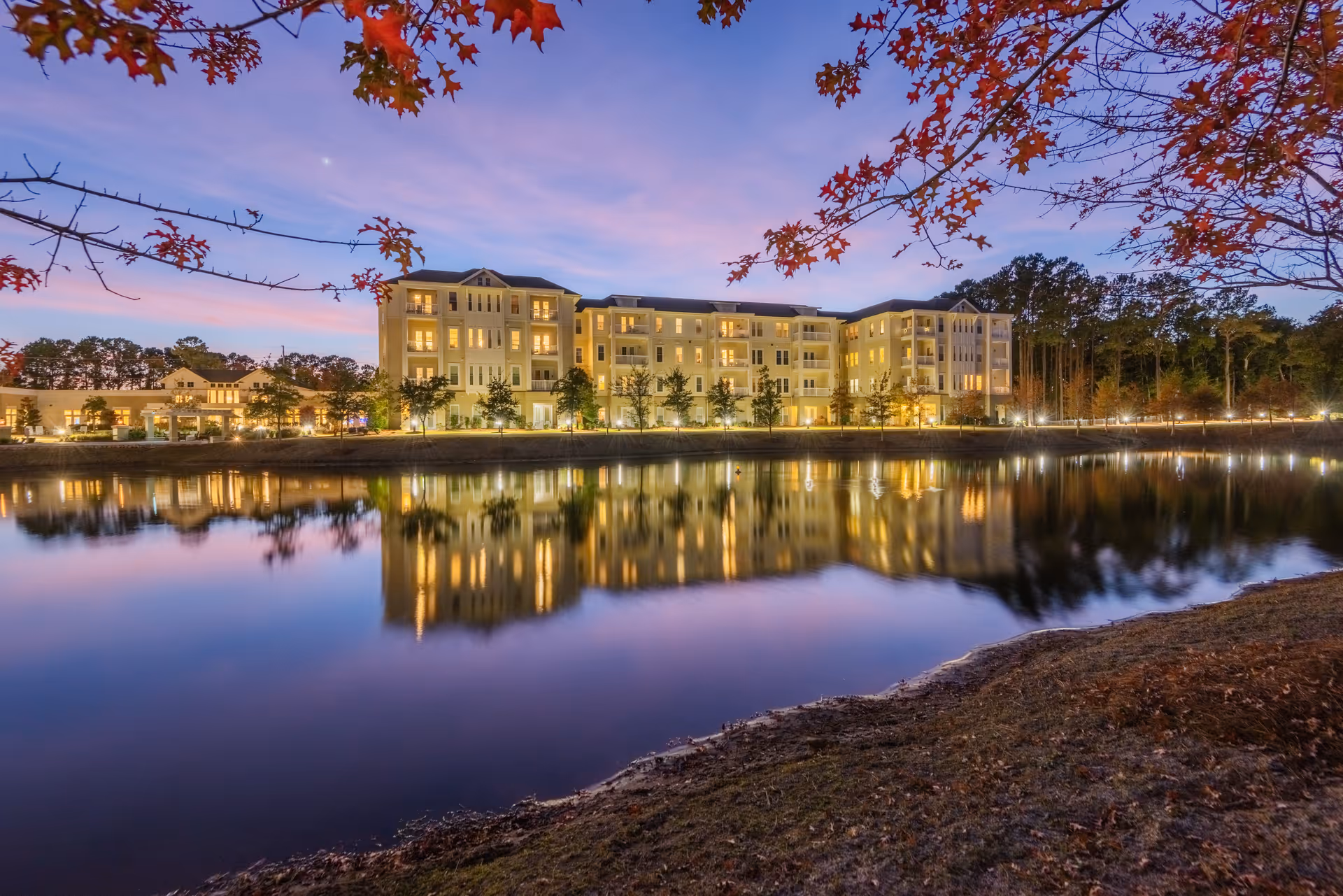 A large, multi-story senior living facility building illuminated at dusk, reflected in a calm body of water in the foreground. Trees with autumn leaves frame the top of the image, and the sky shows a gradient of purple and pink hues.