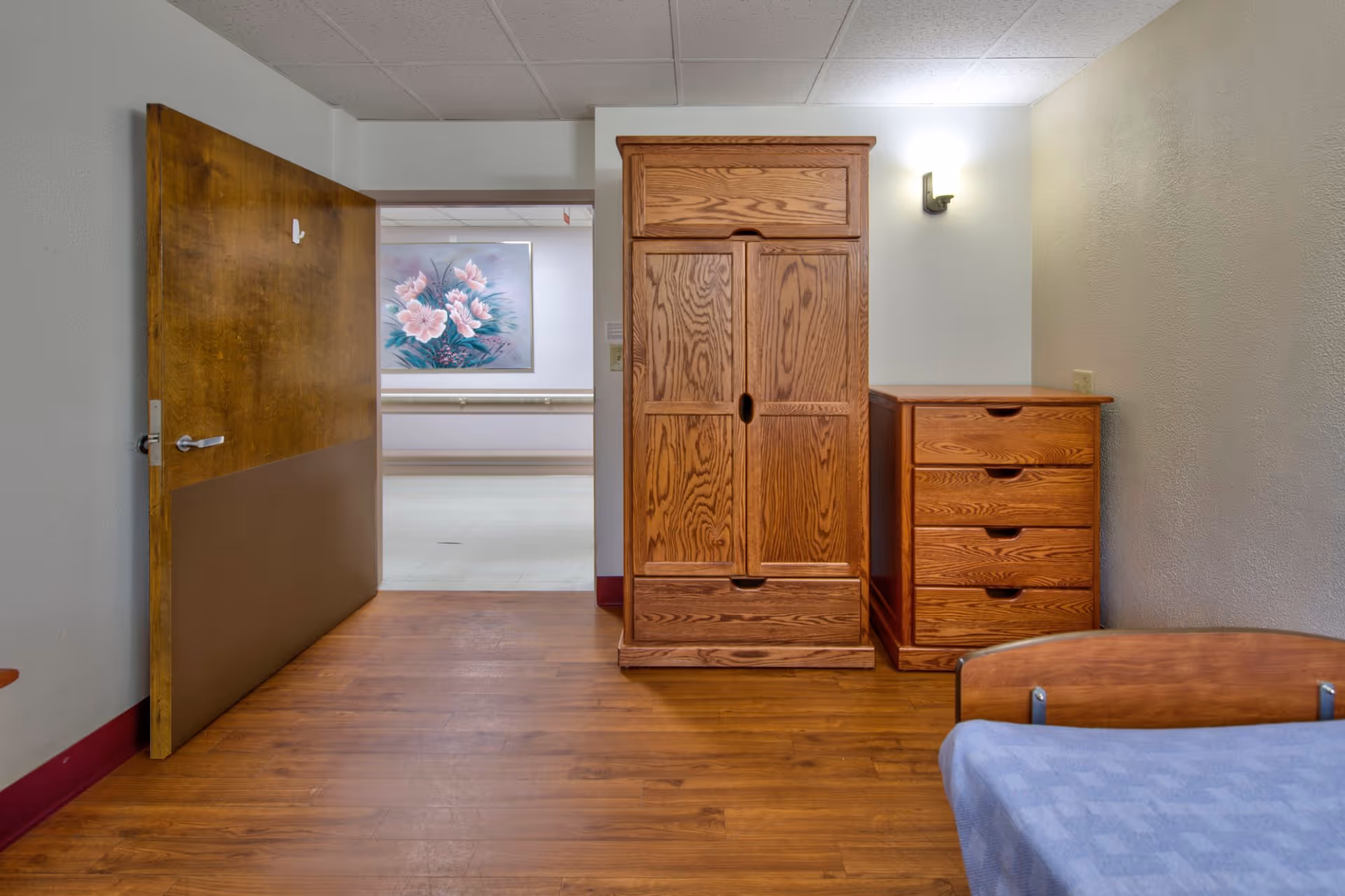 Small senior bedroom featuring a wooden wardrobe and dresser, a partially visible bed, and an open door to a hallway.