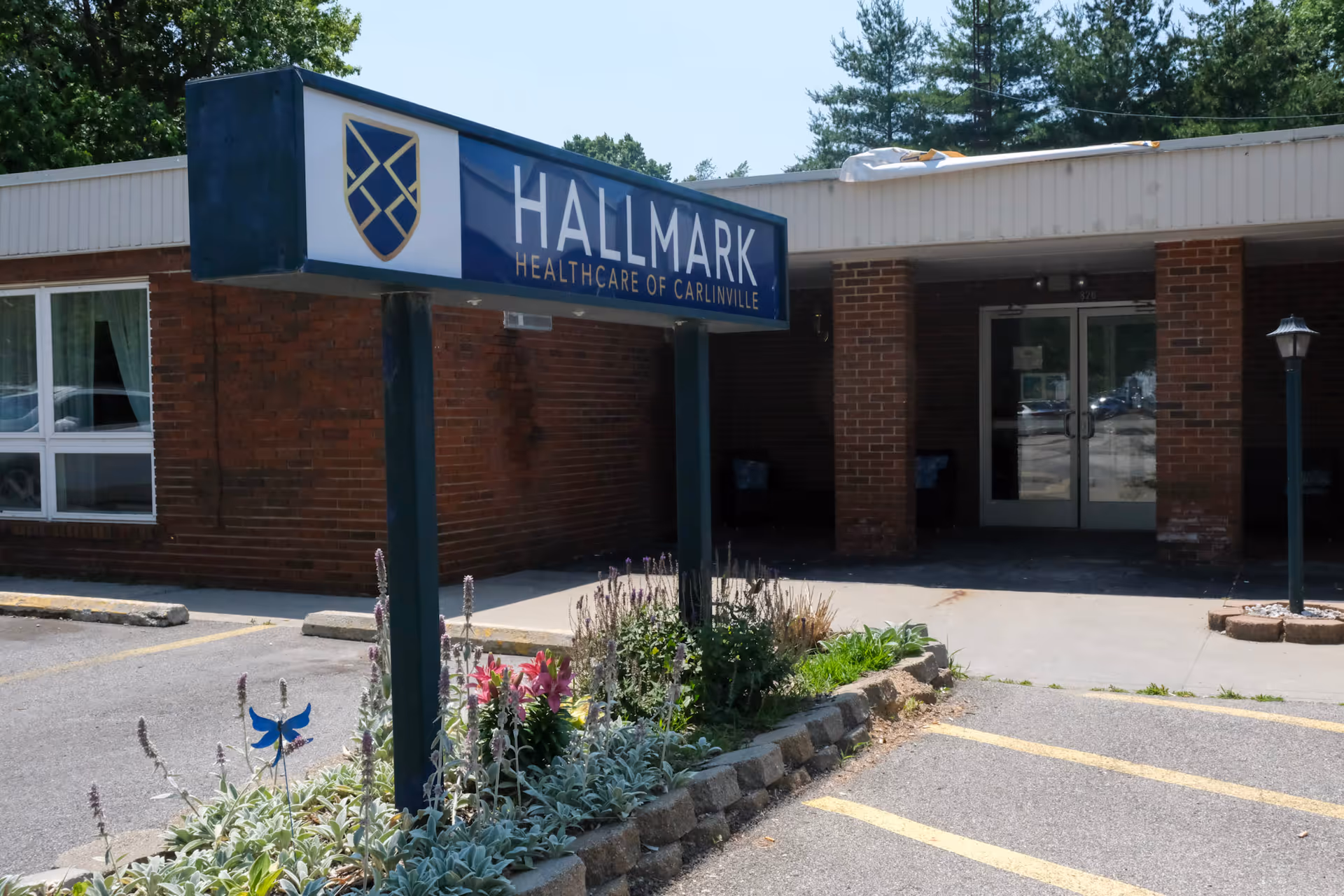 Outdoor entrance showing a Hallmark Healthcare of Carlinville sign in front of a low brick building with landscaping and a driveway.