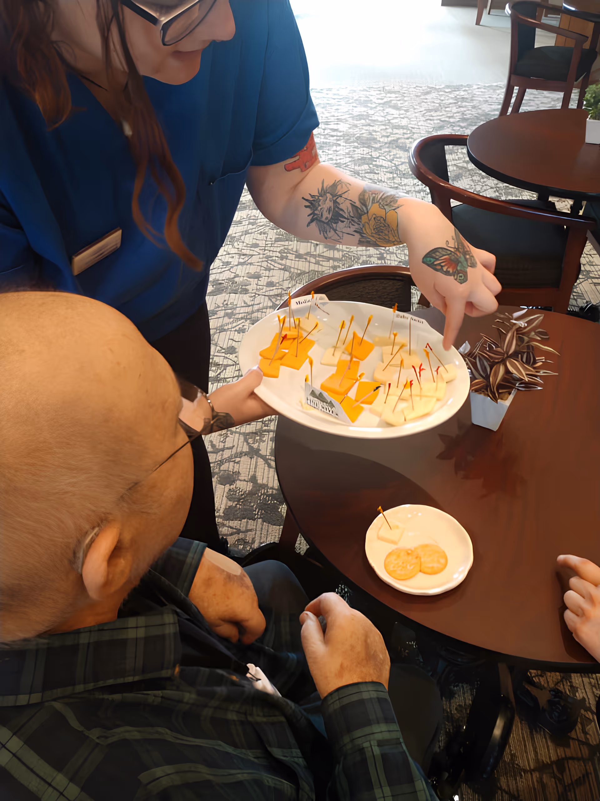 A staff member with tattoos on their arm and wearing glasses offers a plate of cheese samples with toothpicks to an elderly man seated at a table in a communal dining area. The table has a small plate with crackers and a small potted plant. Other tables and chairs are visible in the background.
