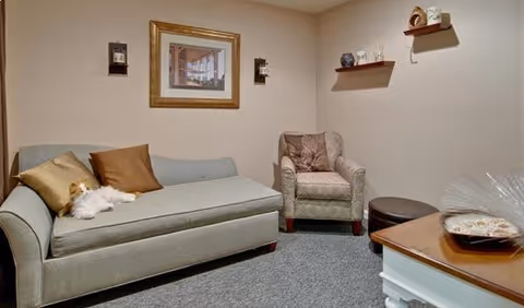 A cozy living room with a light gray sofa adorned with brown and beige pillows and a small white dog resting on it. There is a patterned armchair next to the sofa, a round brown ottoman, and a wooden coffee table with a decorative item on top. The walls are beige with two small shelves holding decorative pieces and a framed picture hanging above the sofa.