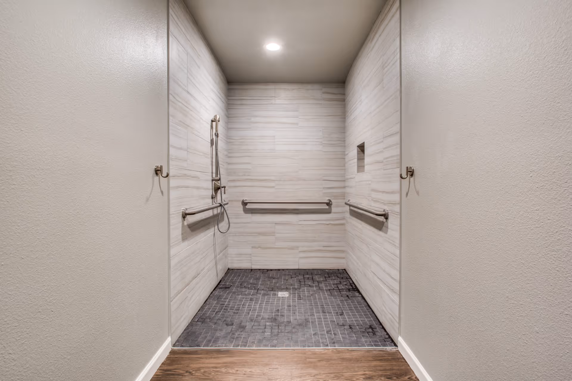 A modern walk-in shower with light beige tiled walls and dark gray tiled floor. The shower features three stainless steel grab bars, a handheld showerhead, and two wall hooks outside the shower area. The ceiling has a recessed light.