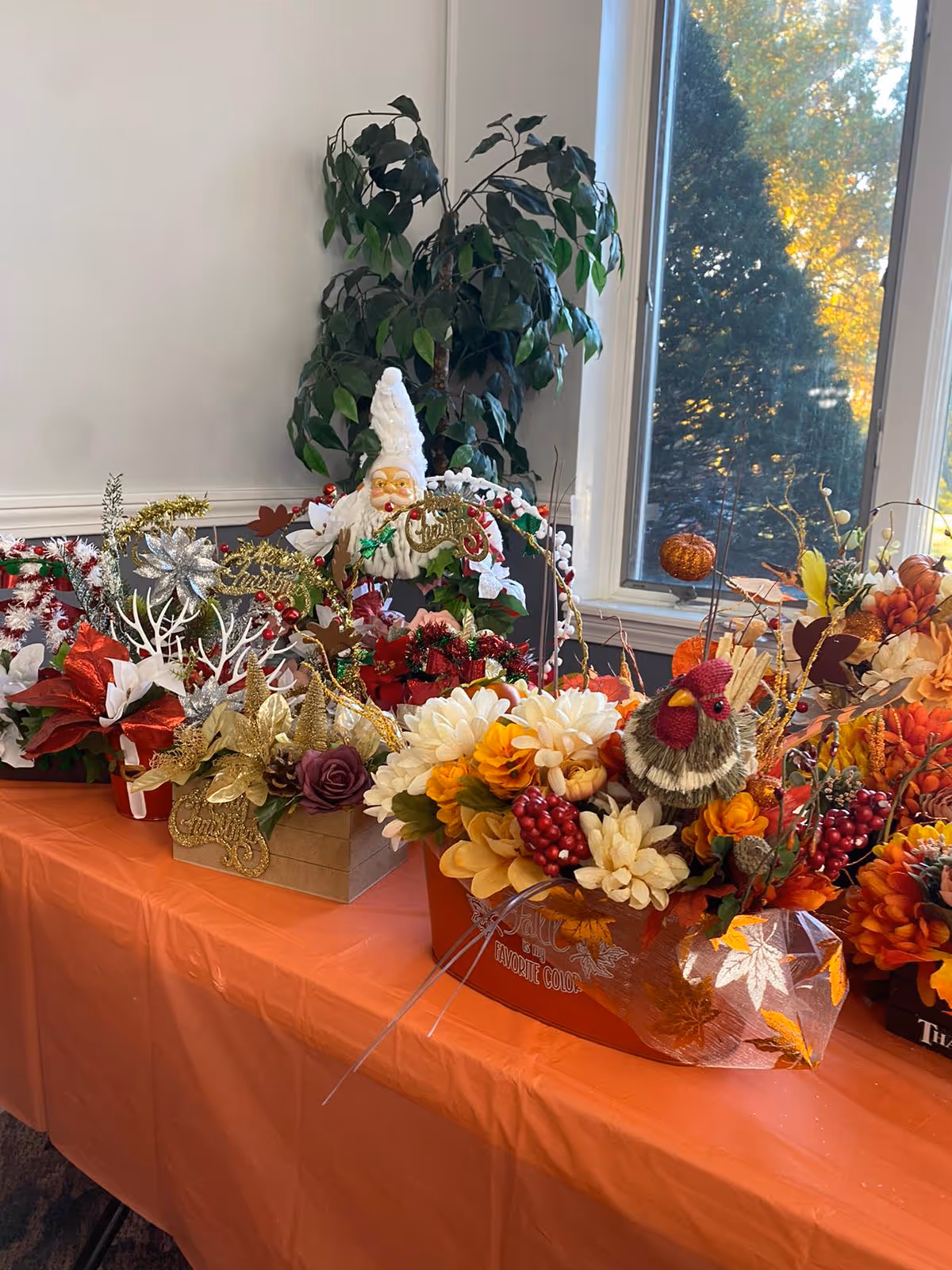 A table covered with an orange tablecloth displaying various festive floral arrangements and decorations, including a Santa Claus figure, autumn-themed flowers, berries, and a small bird ornament. A large green plant is in the background near a window showing trees outside.